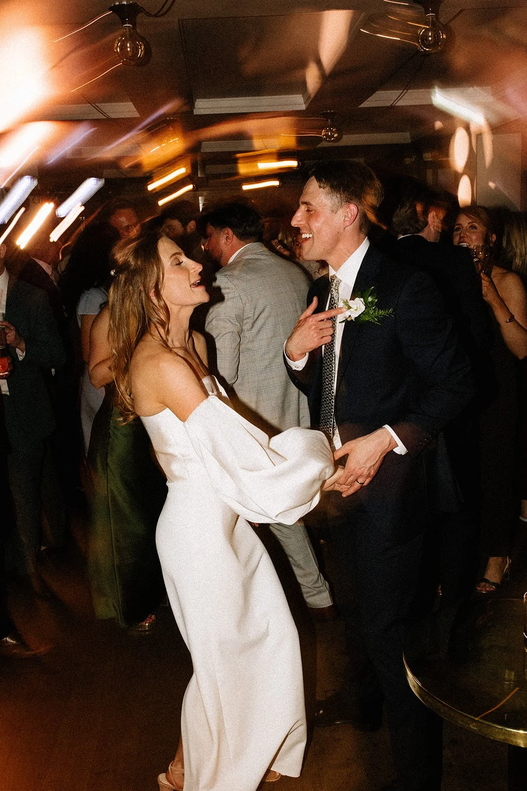 A couple dancing and smiling at a wedding reception, with wedding attire and a lively crowd in the background.