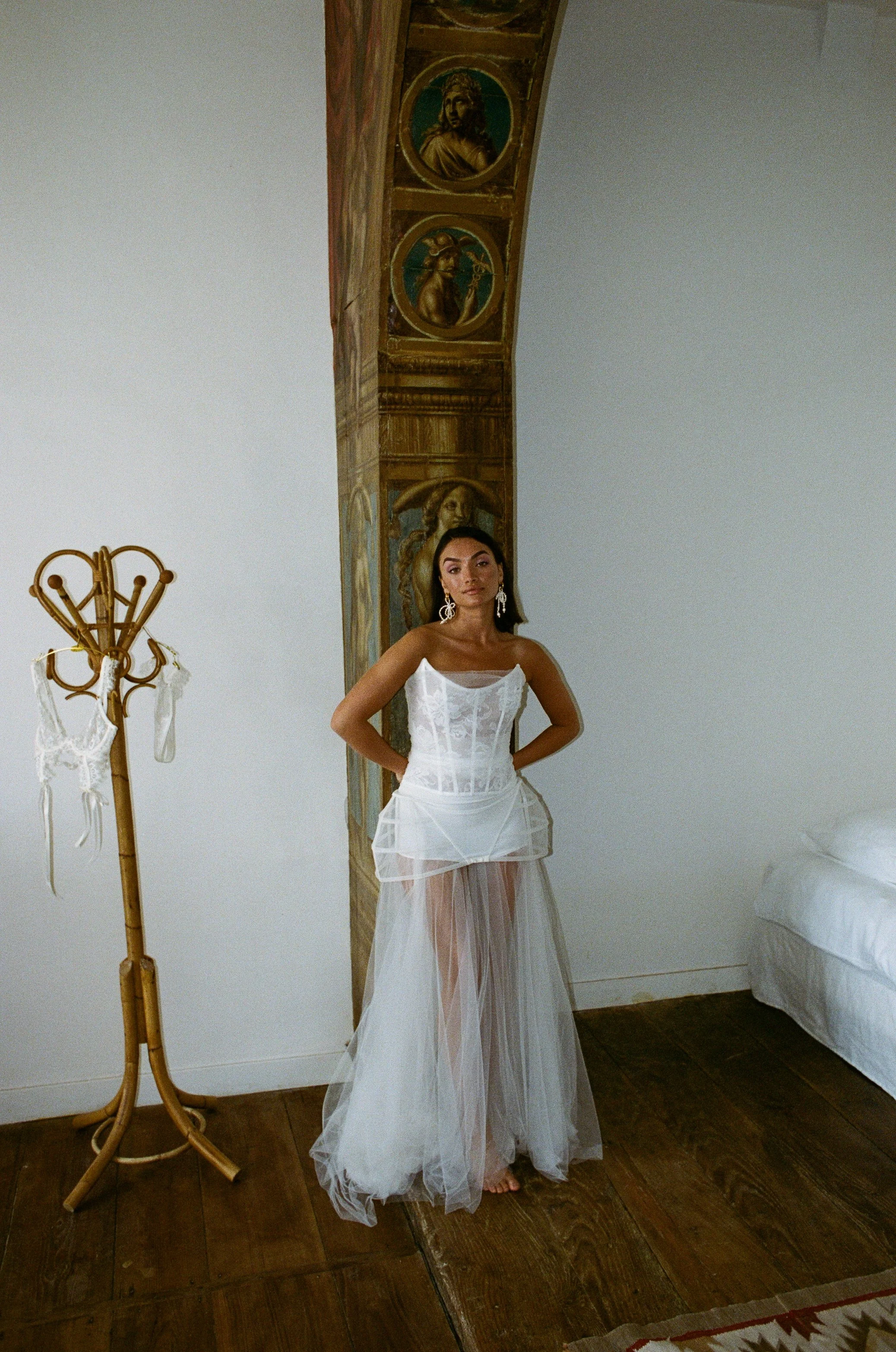 A woman in a white wedding dress with sheer fabric stands in a room with wooden floors and white walls. She has dark hair and is wearing large earrings. There is a coat rack with lace items hanging on it and a wooden strip in the background.