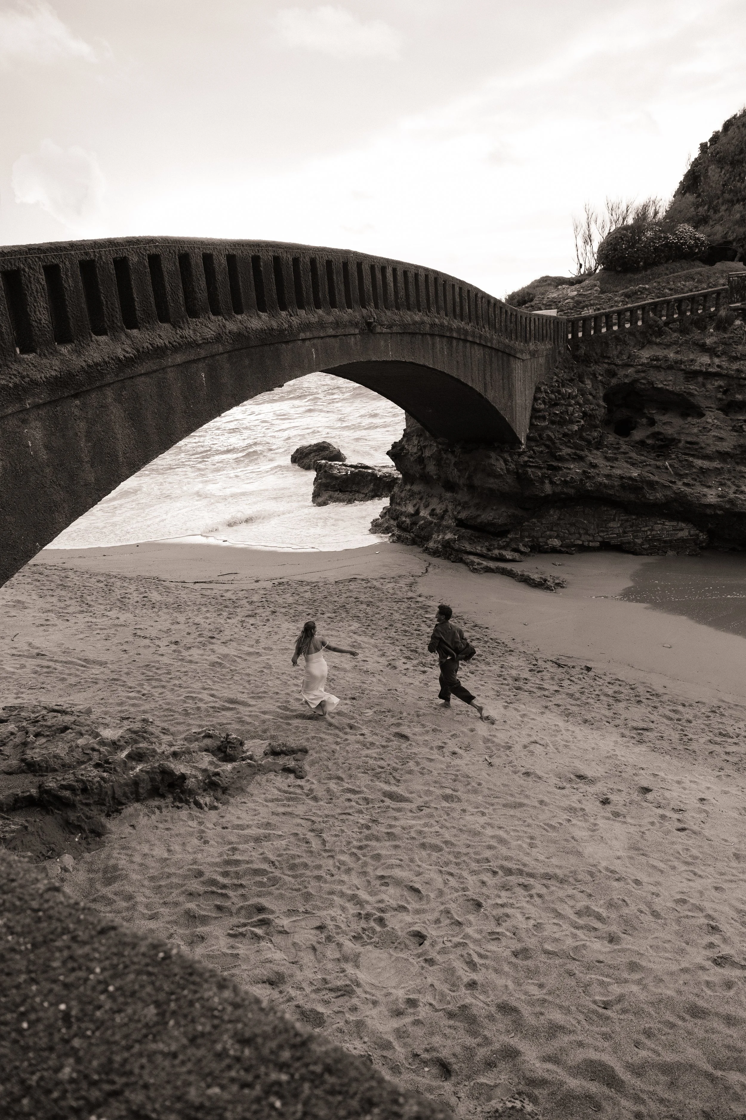 Black and white photo of a large concrete bridge arching over a sandy beach with two people running and playing nearby. The bridge connects rocky cliffs on either side of the shoreline.