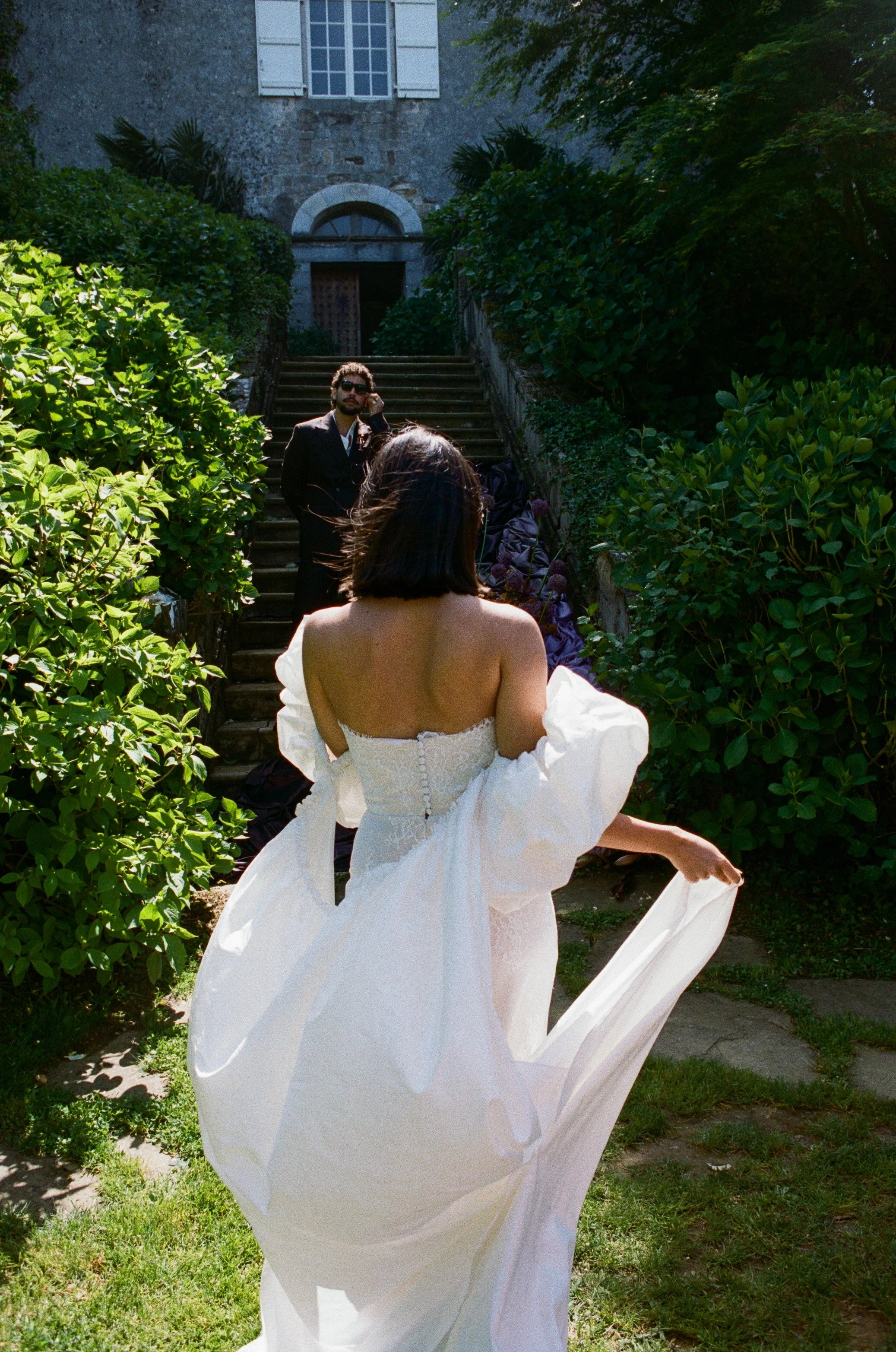 A woman in a white wedding dress is spinning around outdoors on a grassy area, with a man in sunglasses and a black suit standing on stairs behind her, next to a stone building with open windows and greenery.