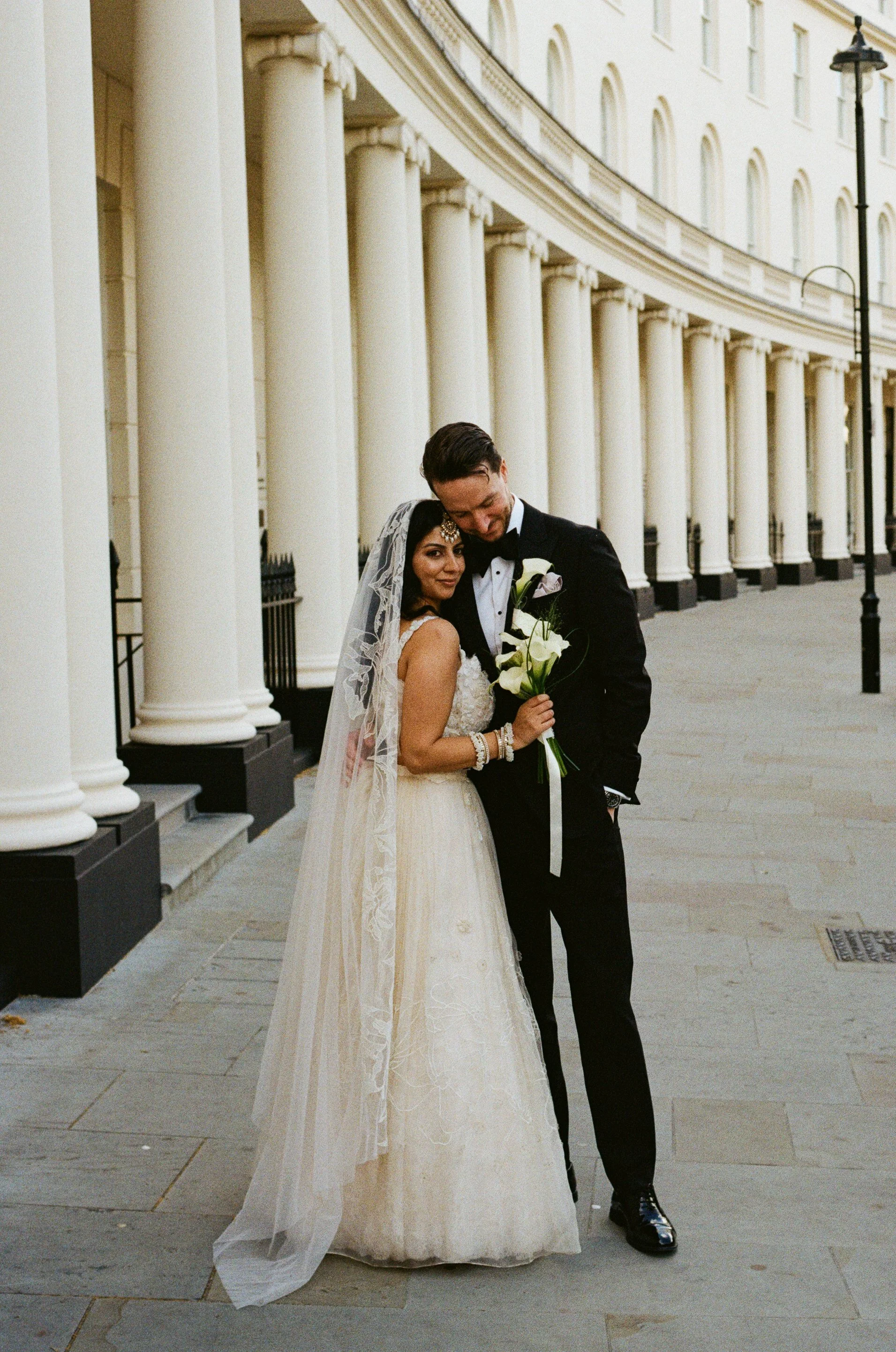 A bride and groom embracing in front of a building with large white columns, the bride holding a bouquet of white calla lilies, both dressed in wedding attire. Taken on 35mm analogue film.