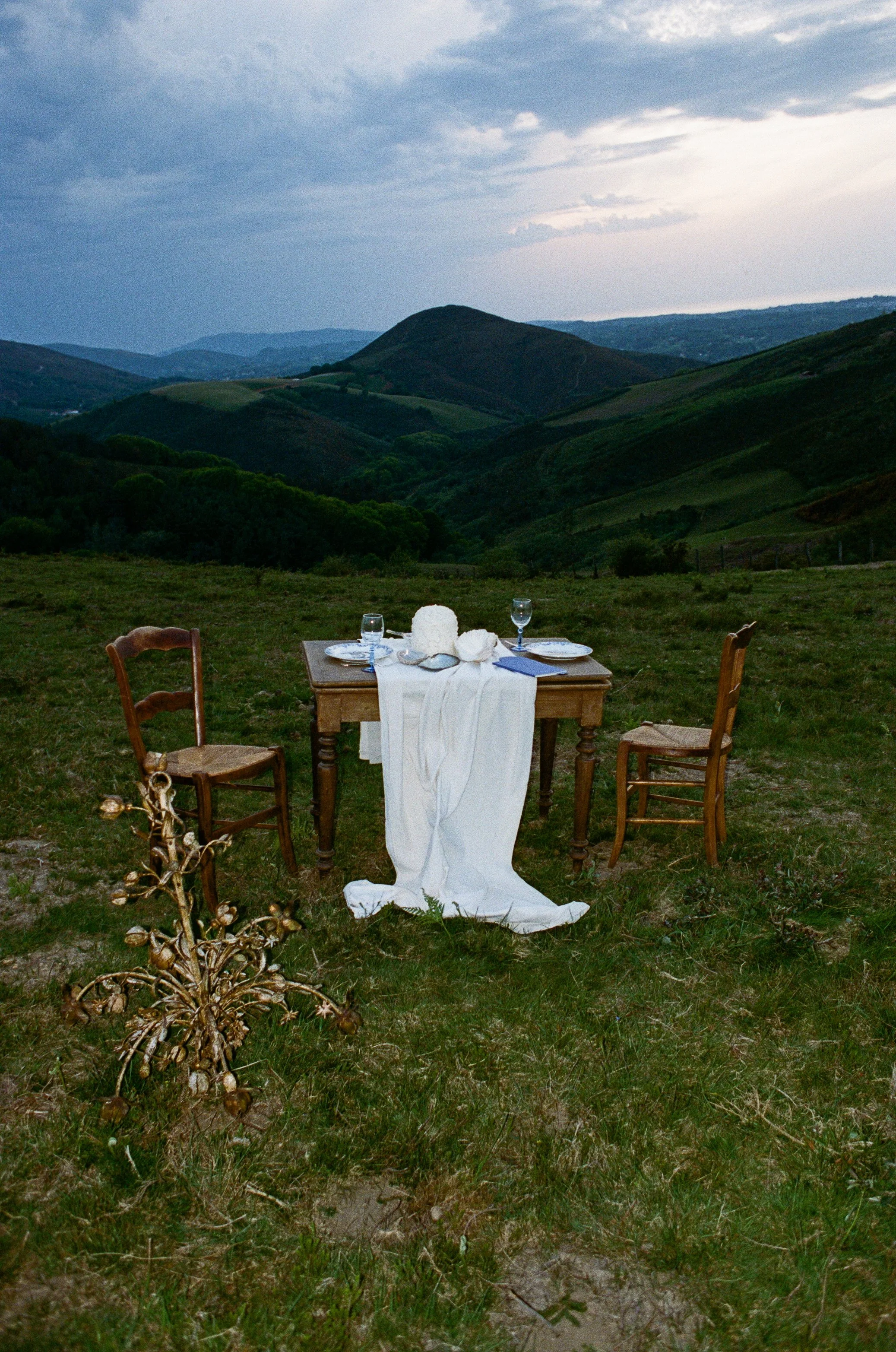A table set for dinner outdoors on a grassy hillside with rolling green hills and cloudy sky in the background.