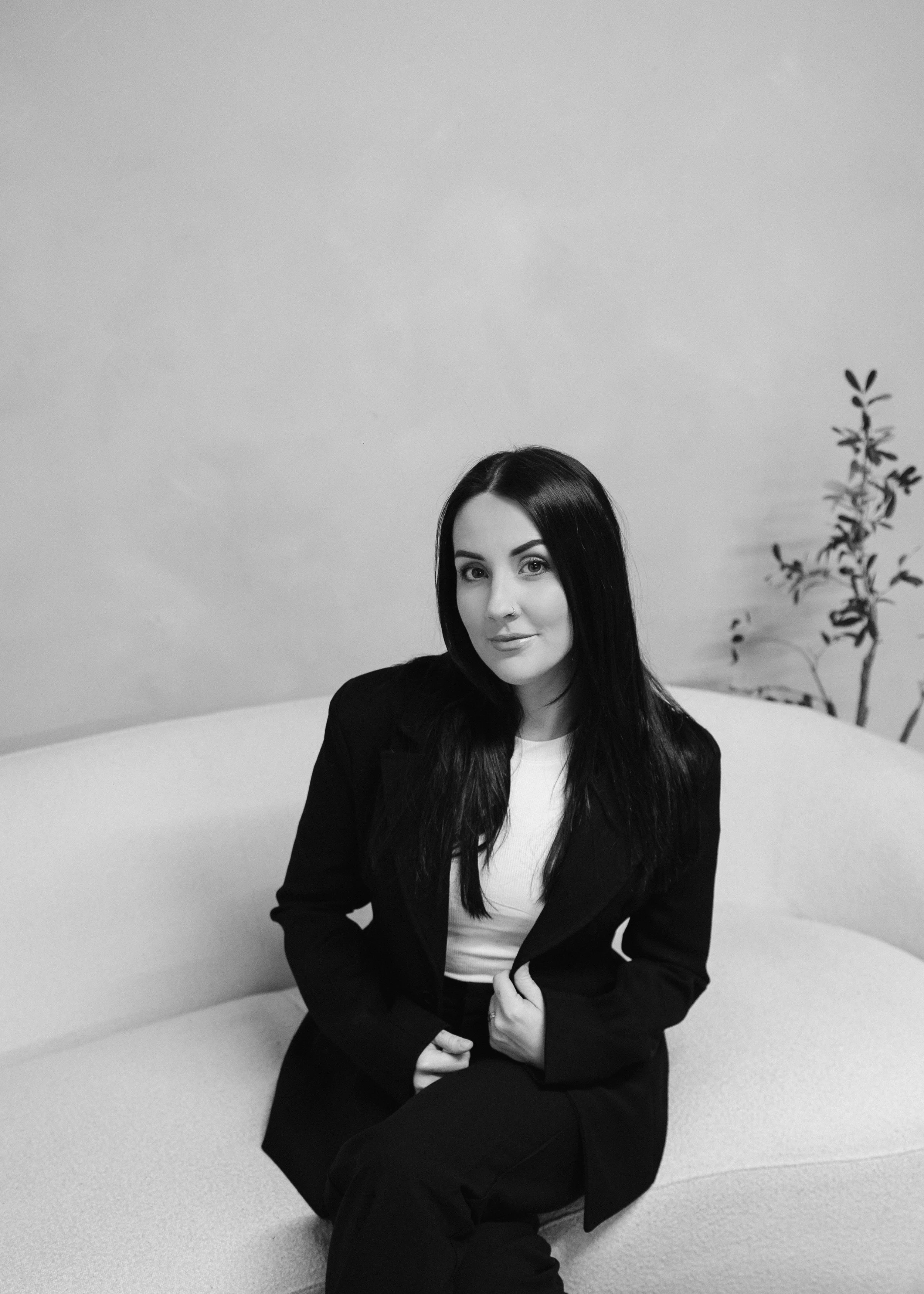 Black and white photo of a woman with long dark hair sitting on a curved sofa, wearing a light-colored top and dark blazer, with a small plant in the background.