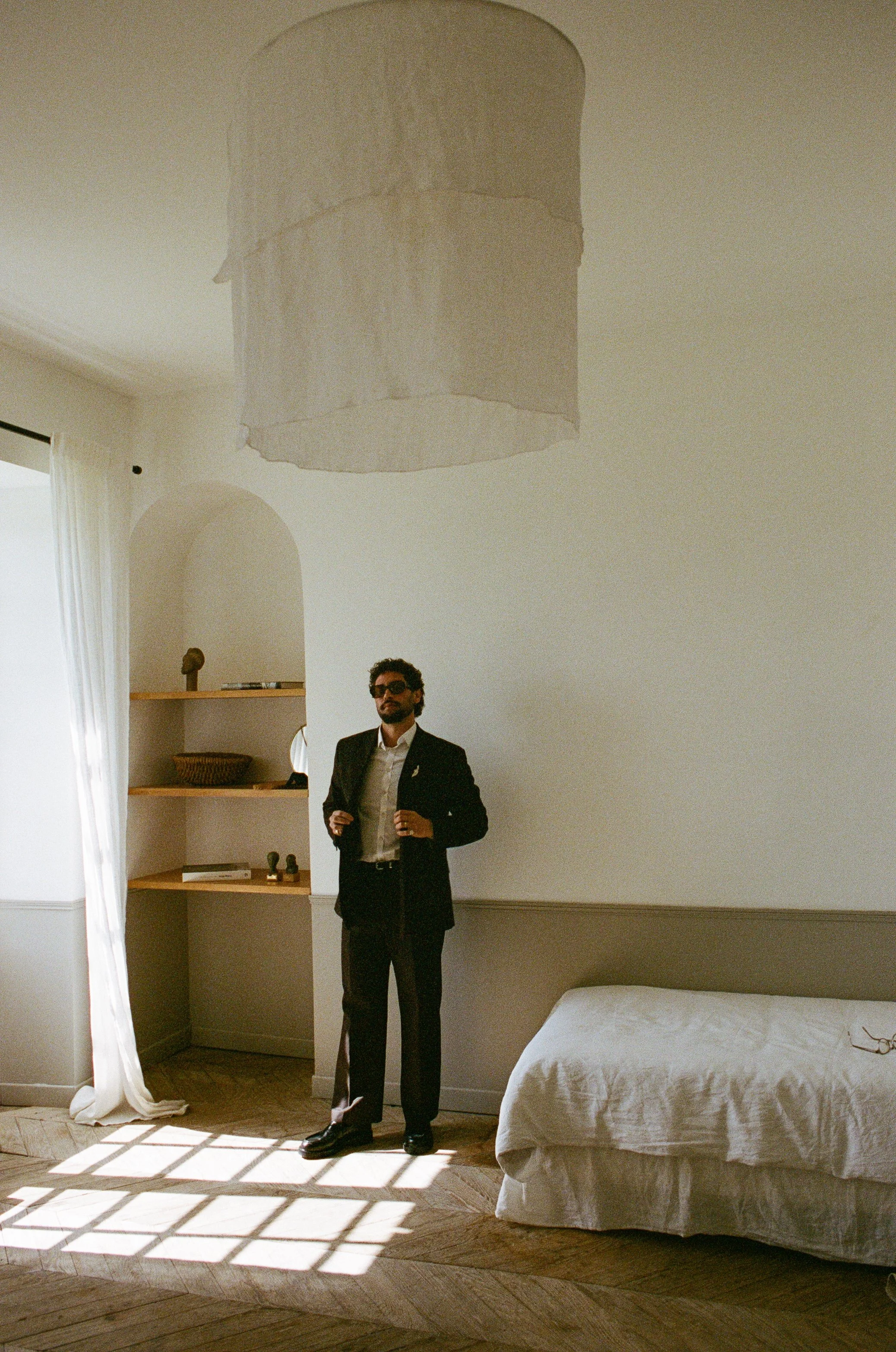 A man in a black suit, white shirt, and sunglasses standing in a sunlit bedroom, with a bed, wooden shelves with decorative items, and a window with curtains in the background. The sunlight creates a checkered shadow on the floor.