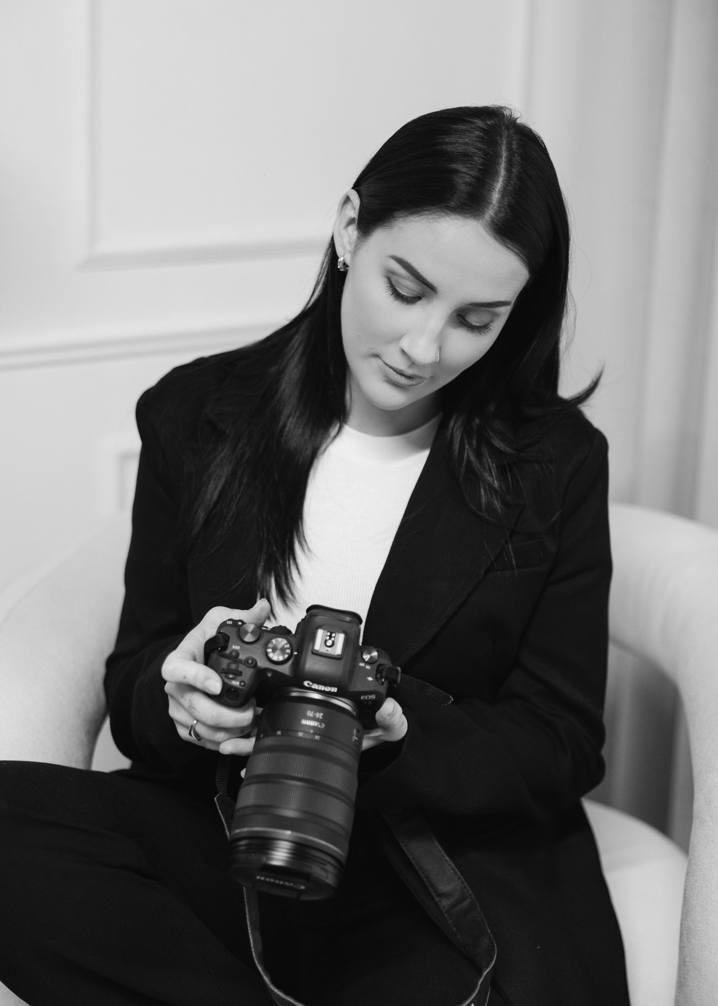 A woman with long black hair and earrings is sitting, looking down at a camera she is holding in her hands. She is wearing a black suit and a white shirt, with a background of white walls, in a black-and-white photograph.