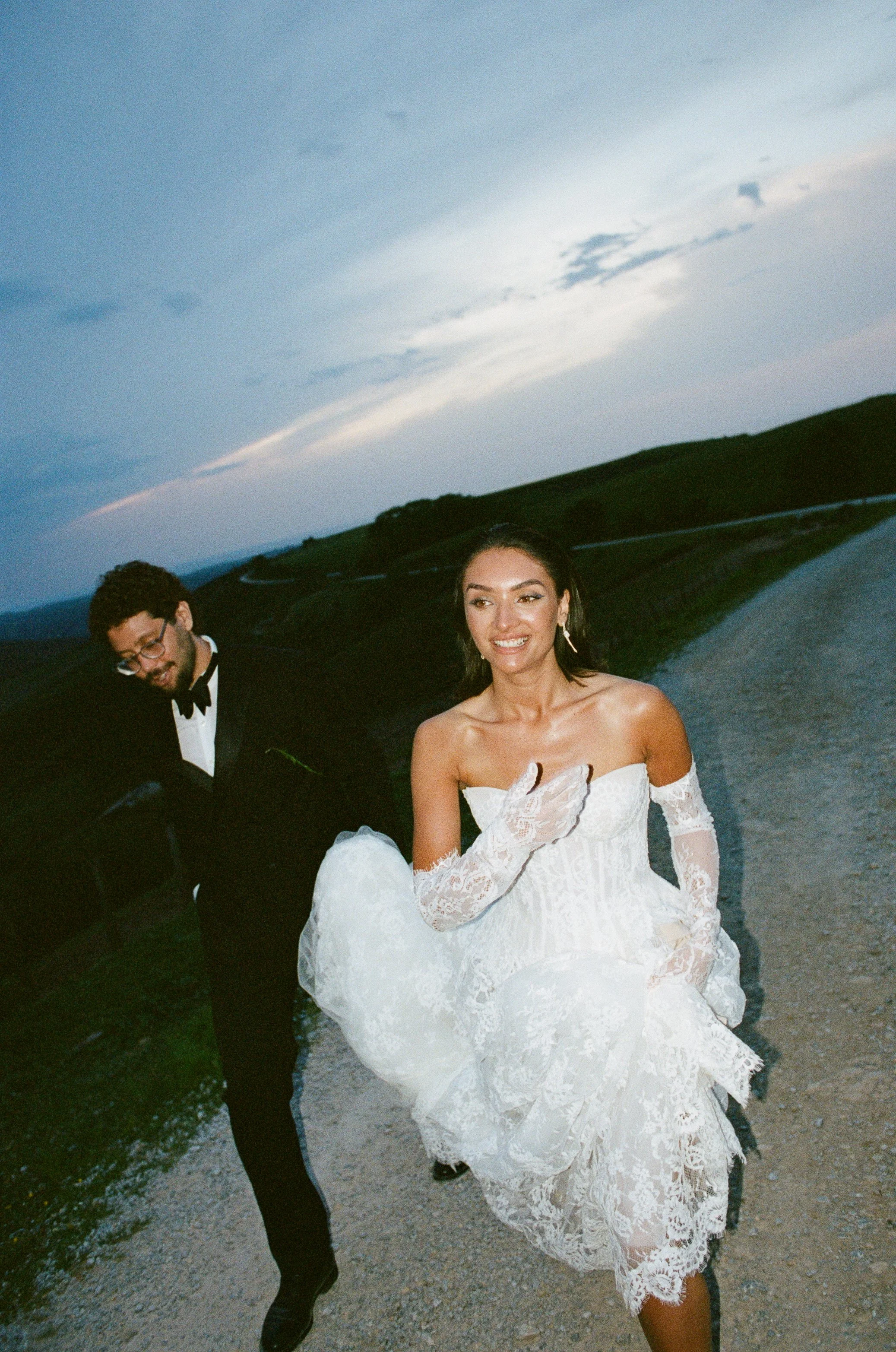 A woman in a white wedding dress walking on a gravel road with a man in a tuxedo and bow tie on her left, in a rural landscape with hills and a cloudy sky.