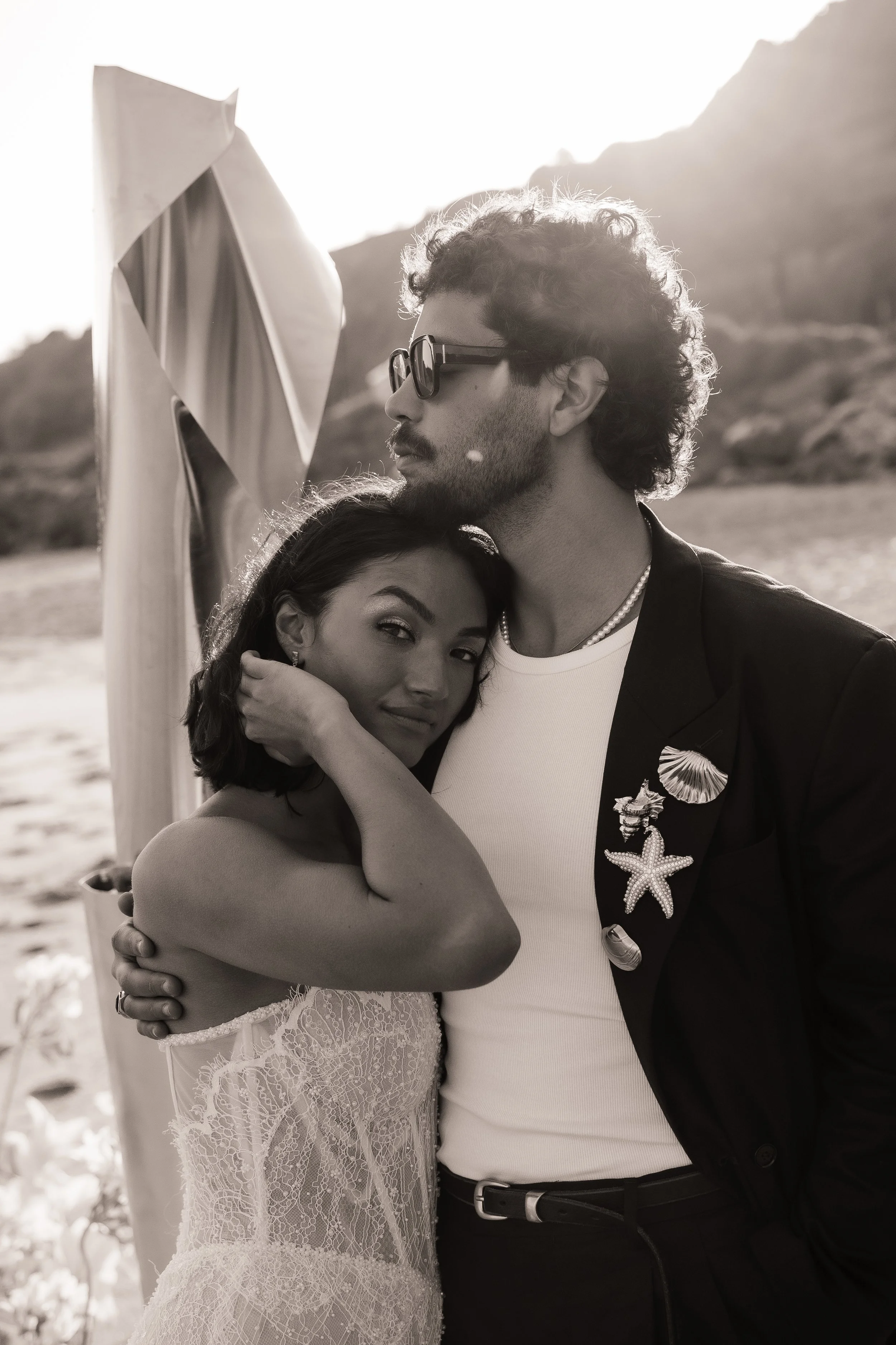 A couple hugs on a beach with a sculpture in the background, the man wearing sunglasses and a blazer with a starfish brooch, the woman in a lace dress, both smiling, black and white photograph.