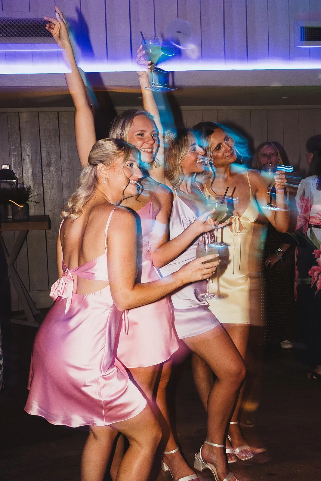 Group of women enjoying a night out at a party, dancing and holding drinks, wearing colorful satin dresses.