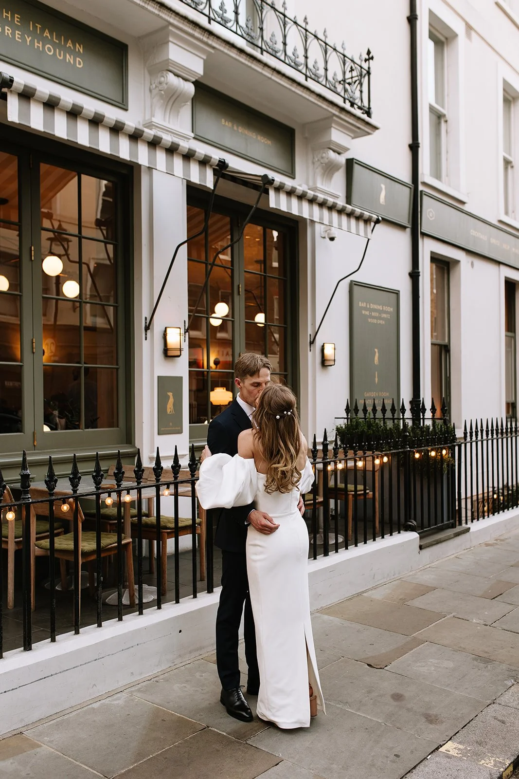 A couple is dancing and kissing outside a restaurant with string lights and black railing, with a city street in the background.