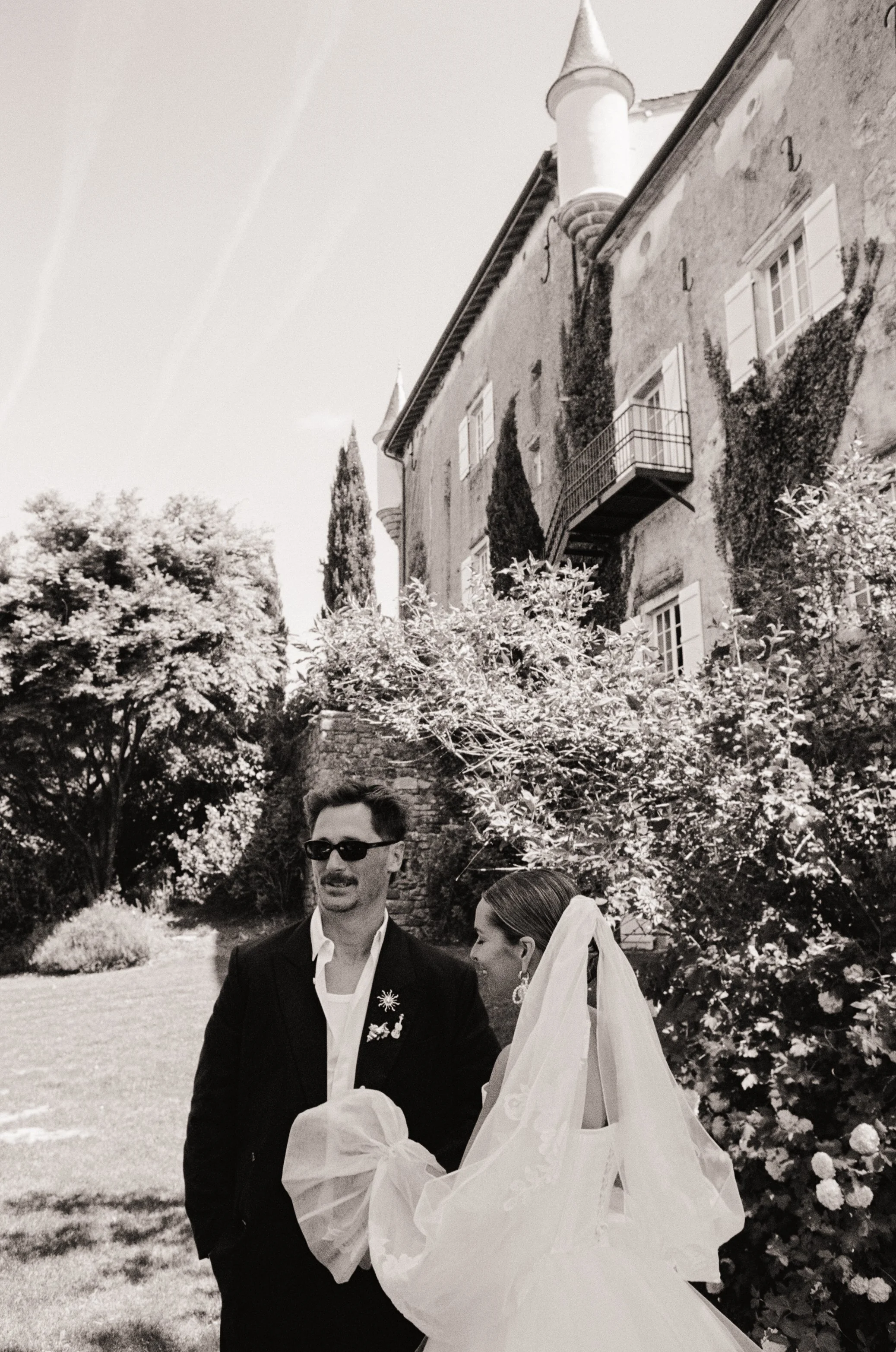 A black and white photo of a man and woman on their wedding day. The man wears sunglasses and a suit, the woman has a veil and earrings. They are outdoors with trees, a stone wall, and a large house with windows and a turret in the background.