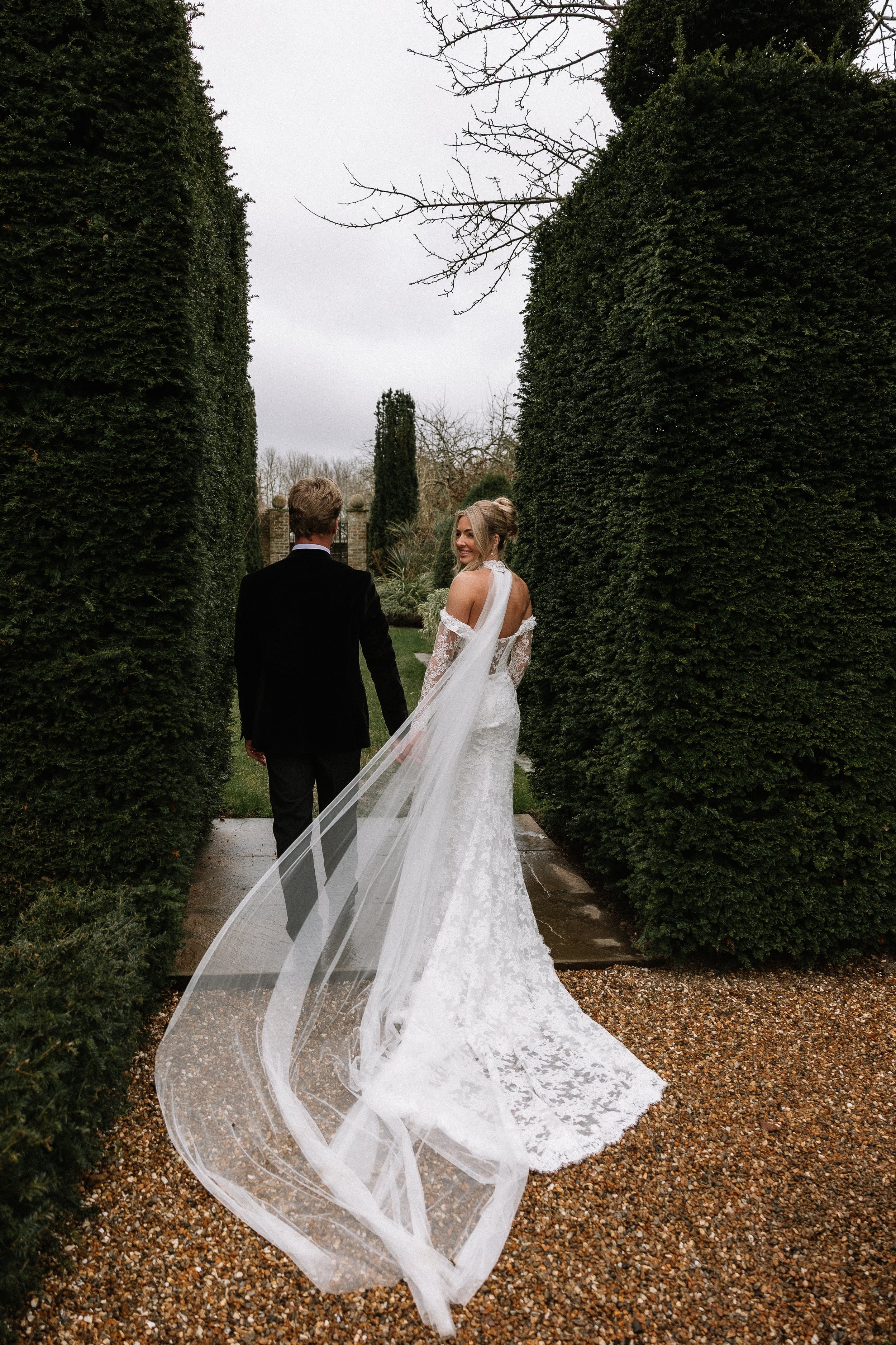 A bride and groom walking hand in hand through a garden pathway lined with tall, green hedges. The bride is in a white lace wedding dress with a long train and a veil flowing behind her. The groom is in a black tuxedo. The sky is overcast.