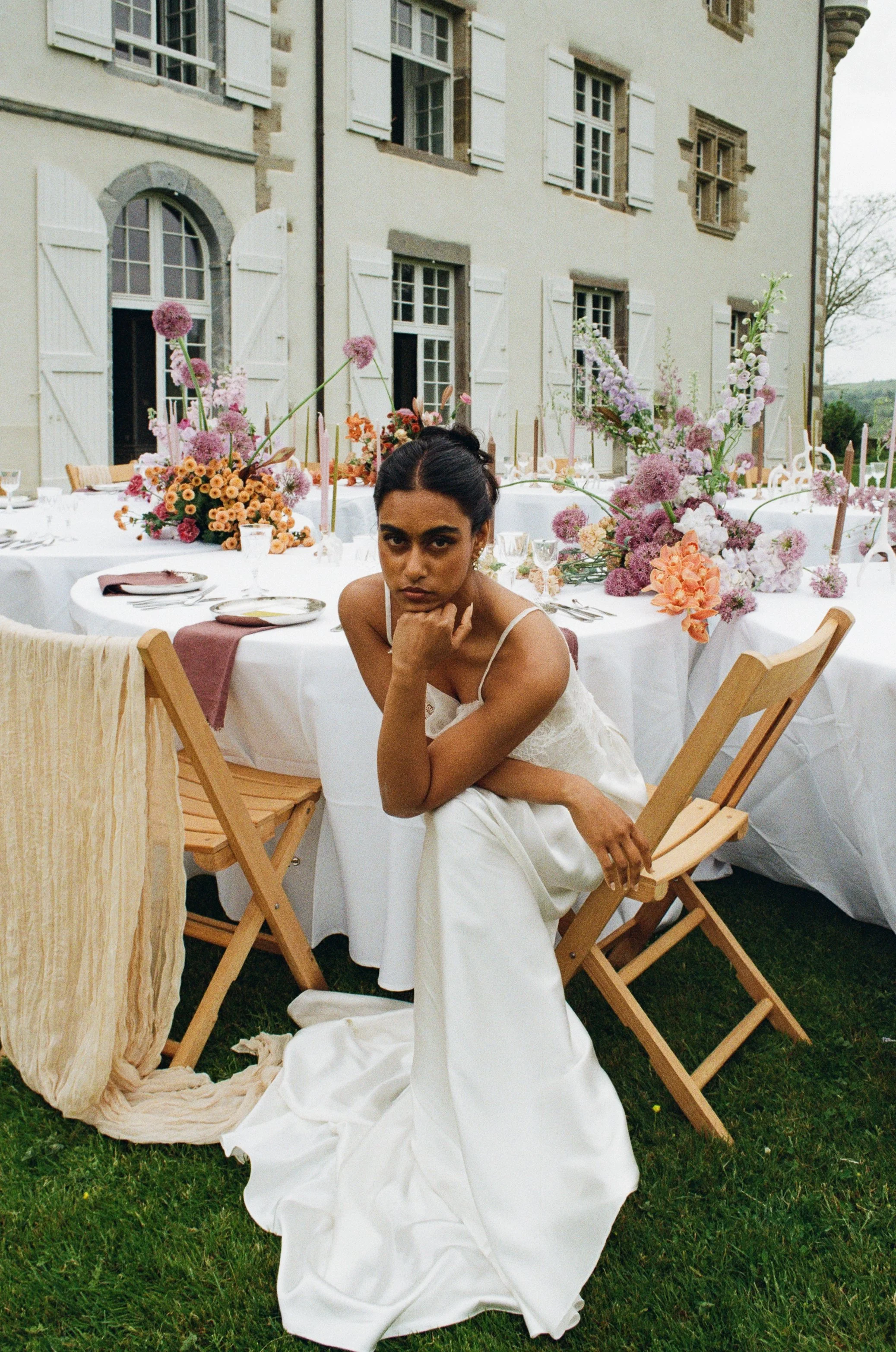 A woman in a white dress sitting on a chair outdoors at a decorated event with tables, flowers, and a historic building in the background.
