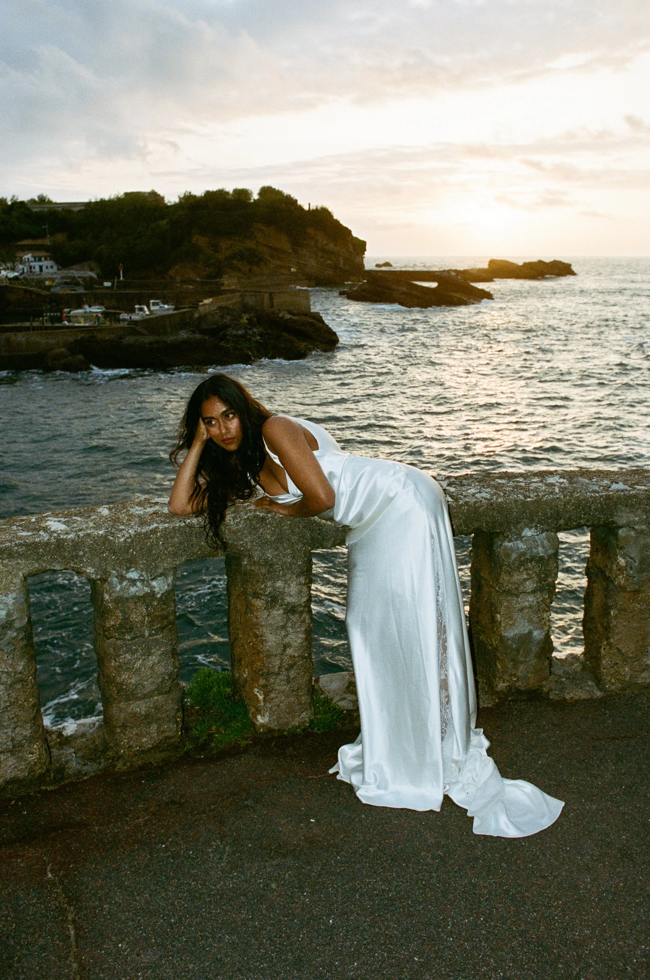 A woman in a white dress laying on a stone railing by the ocean at sunset.