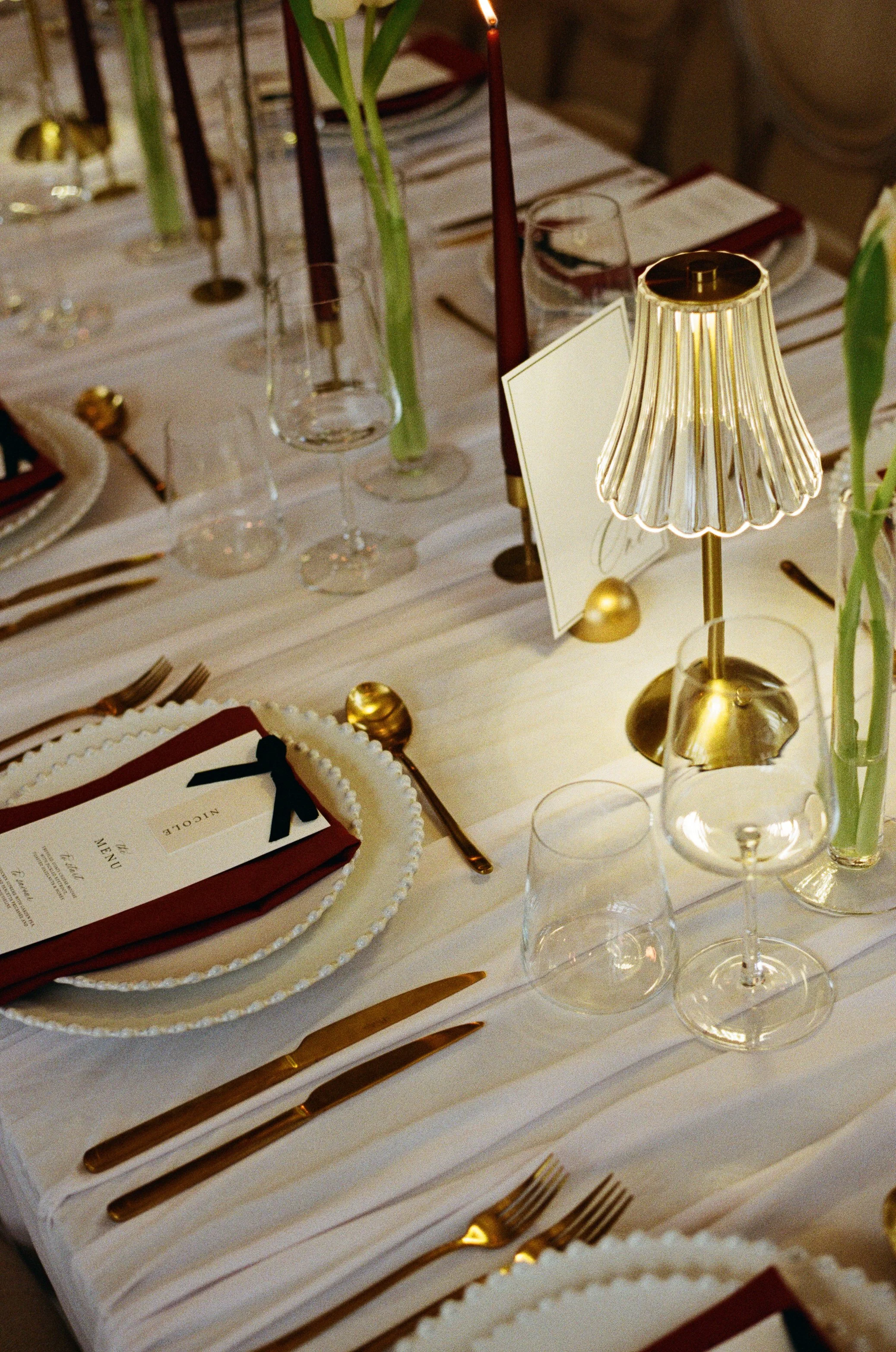Elegant table setting with gold-plated utensils, wine glasses, and a menu. A small gold lamp provides warm lighting. Centerpieces include tall green flowers in glass vases and red candles. The table is decorated with gold accents and a white tablecloth.