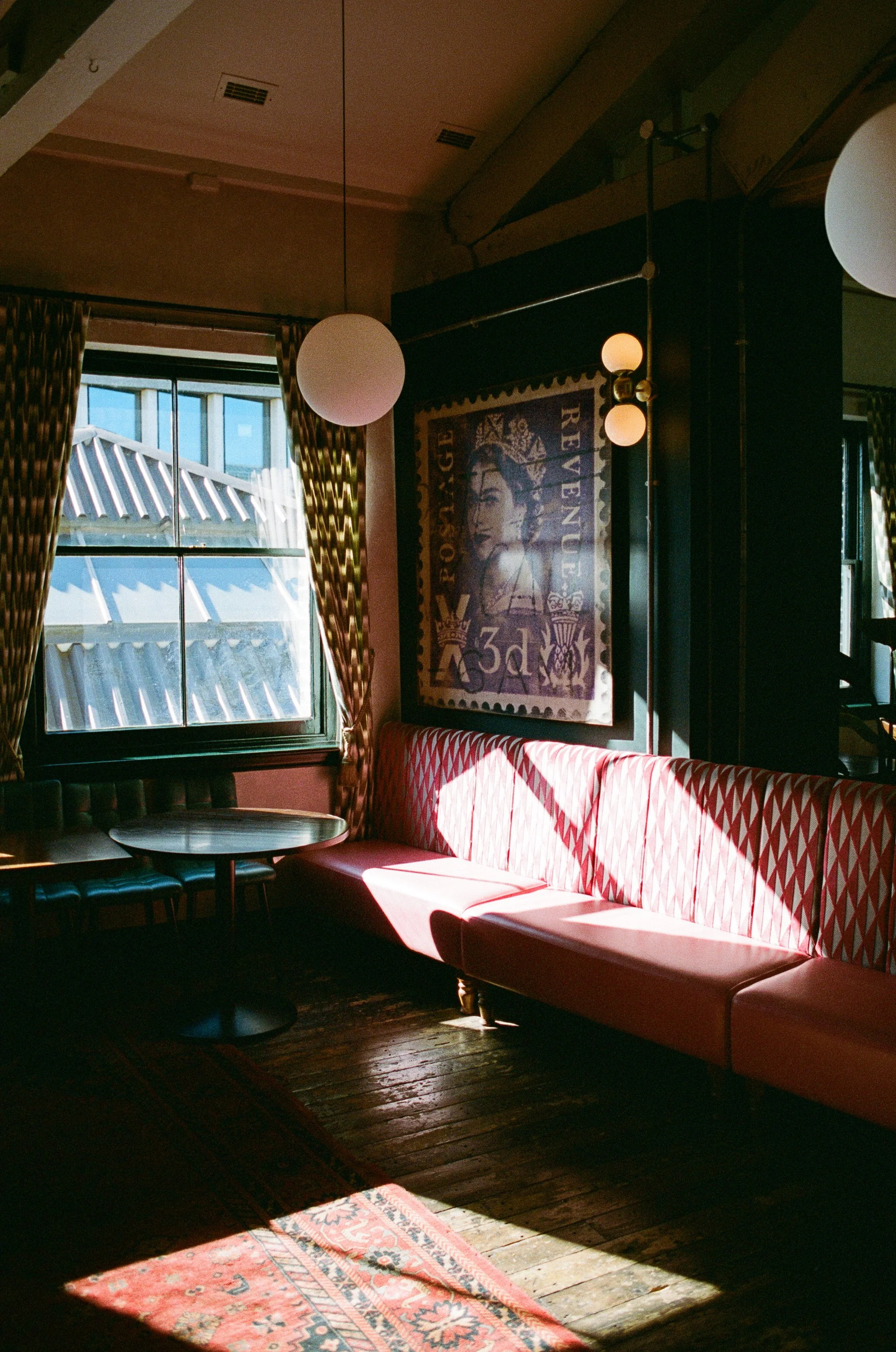 Interior of a cozy room with a pink upholstered bench seating along the wall, a round table, and a window with patterned curtains. Sunlight creates a warm glow on the wooden floor and a decorative rug. There is a framed artwork on the wall.