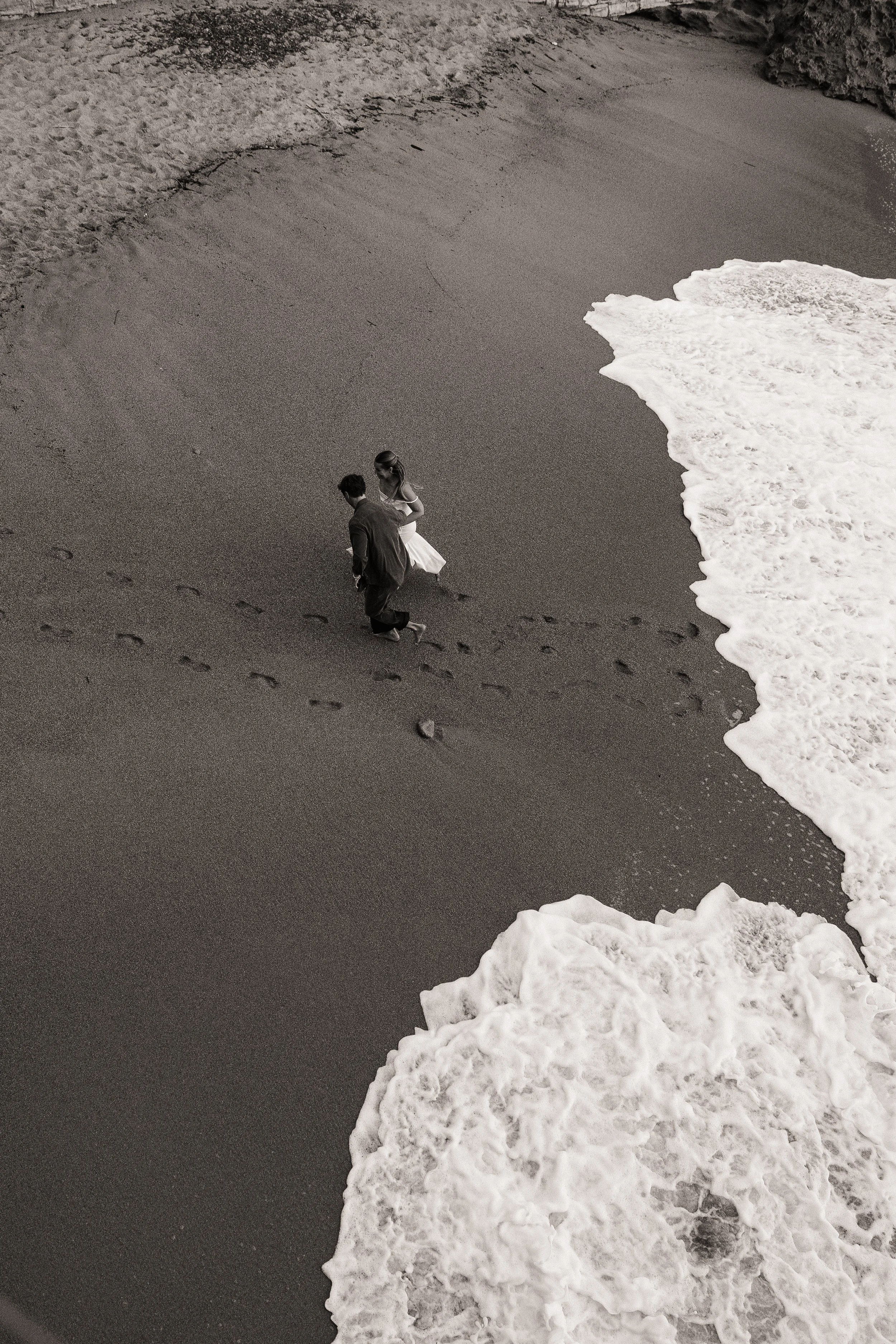 Two people walking along a sandy beach near the water's edge, with snow or ice piled along the shoreline, in black and white.