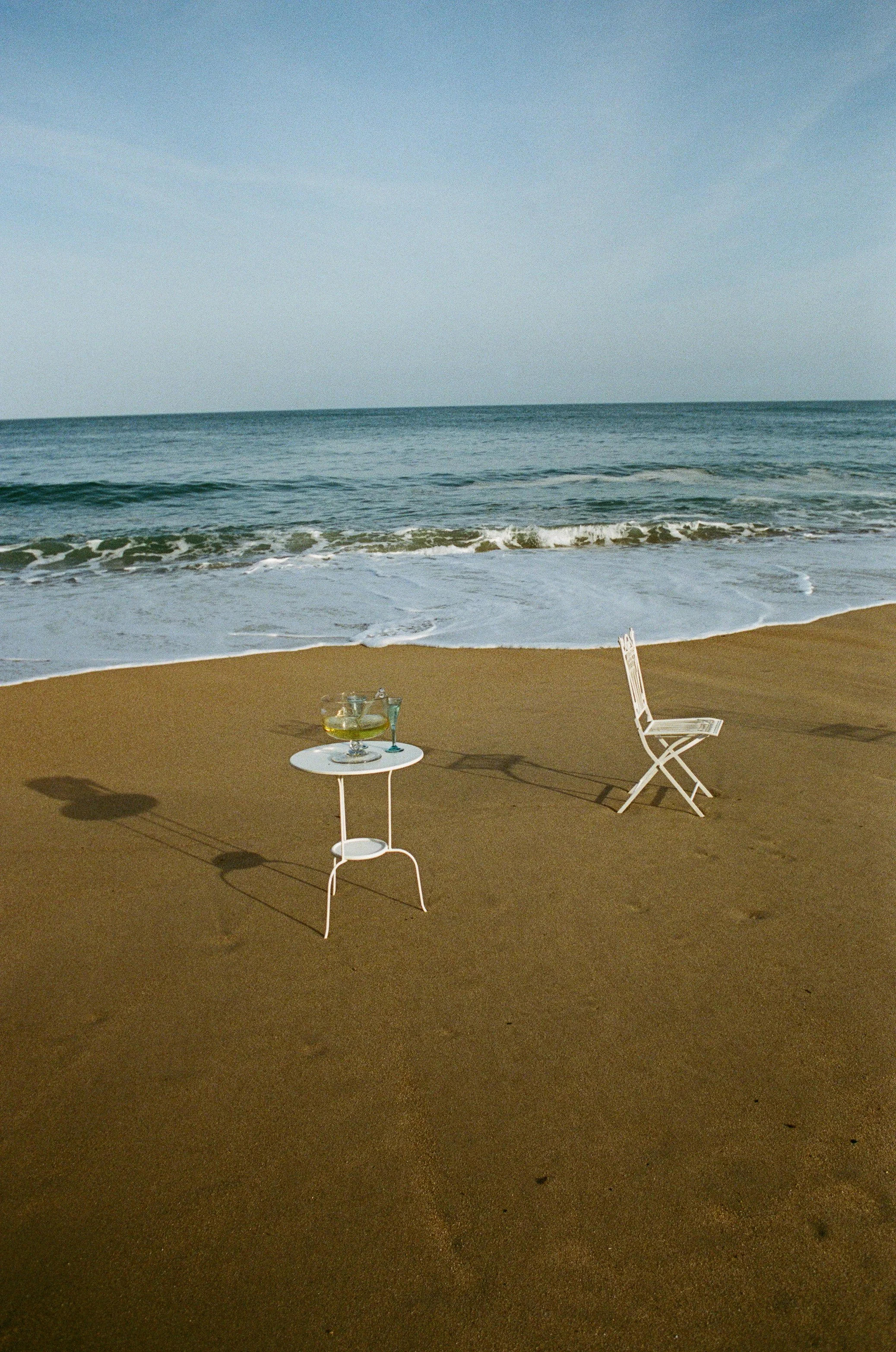 Empty beach with a small white table holding two glasses of drinks and a white chair nearby, with the ocean and sky in the background.