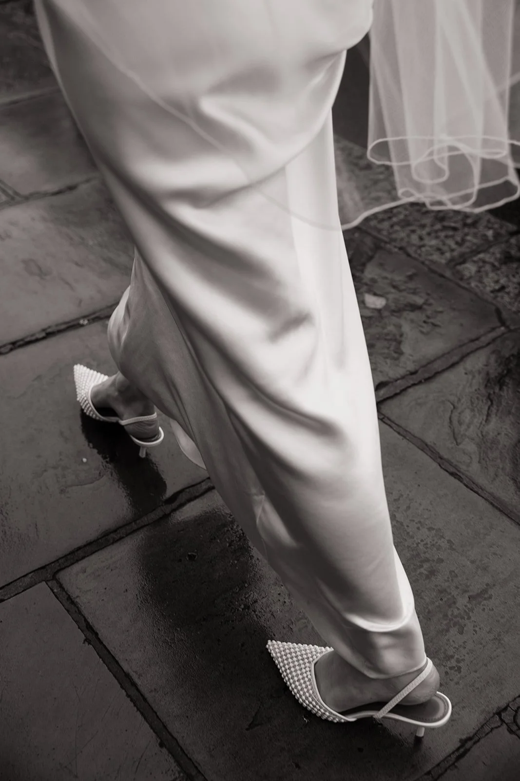 Close-up of a person wearing a long satin or silk gown, elegant high-heeled shoes, and a veil, standing on wet stone pavement.