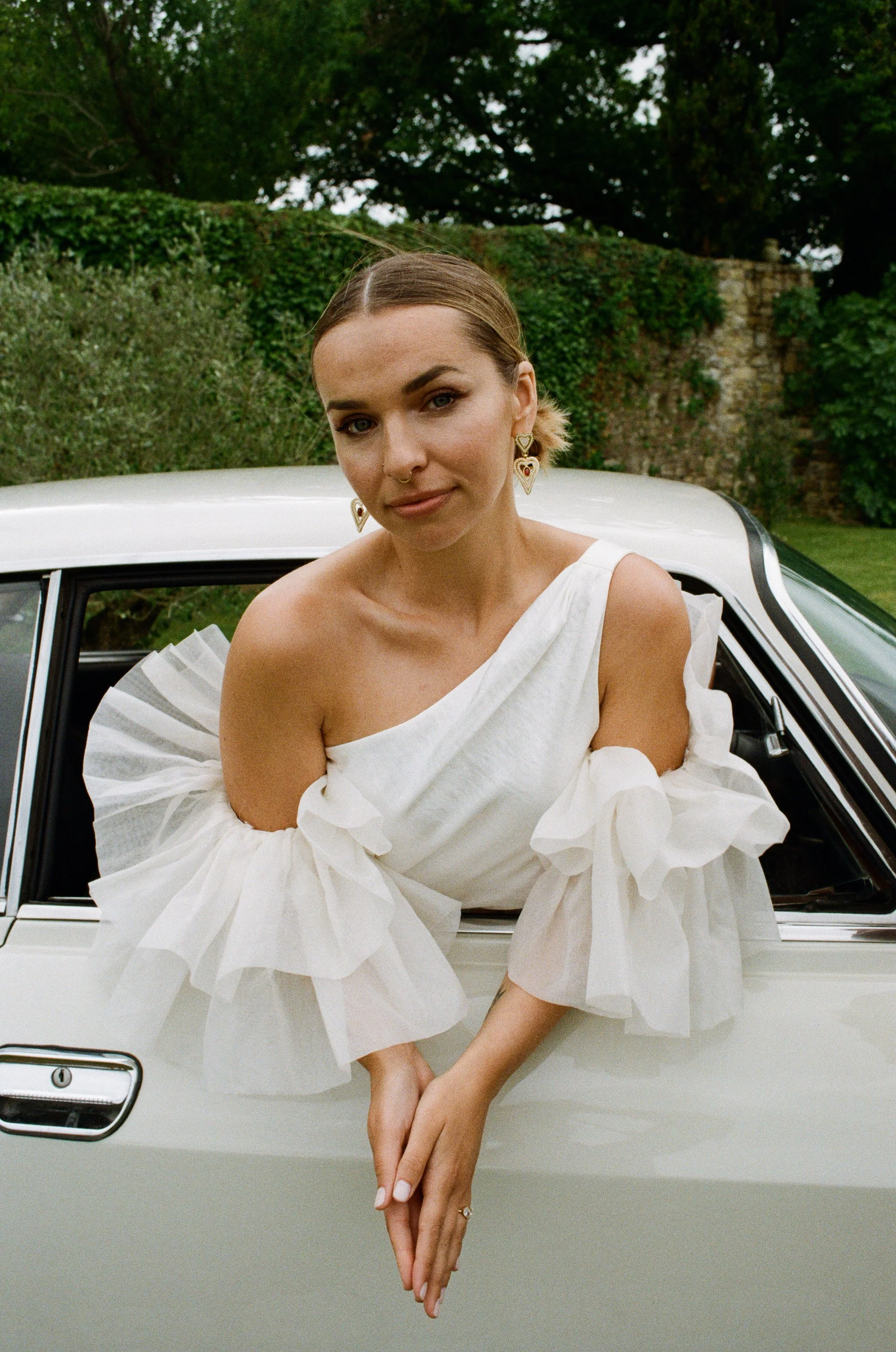 A woman with short brown hair and jewelry, wearing a white dress with puffed sleeves, sitting on the door of a white vintage car in a garden with greenery and trees in the background. Taken on 35mm analogue film.