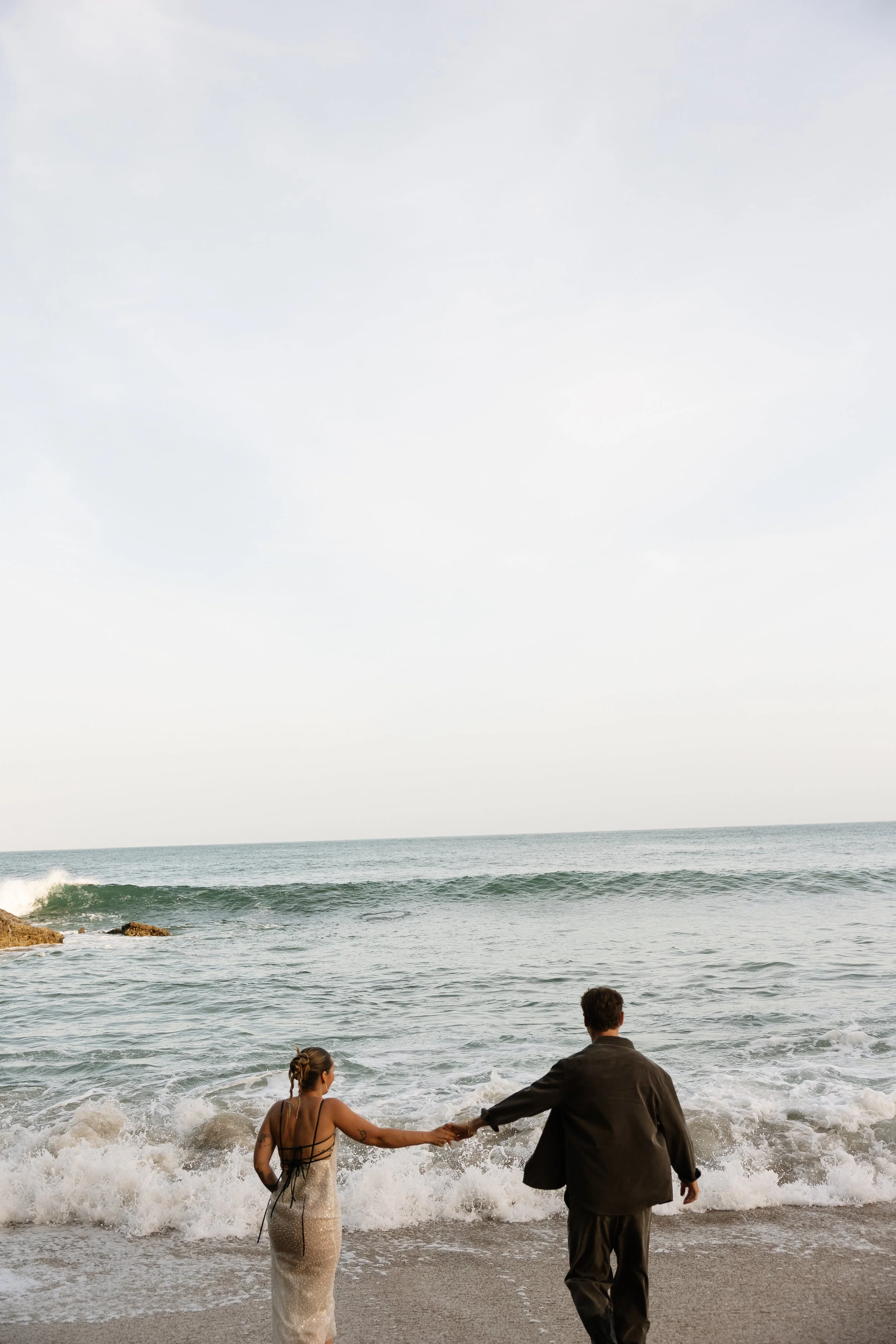 A couple holding hands at the beach, with the woman in a beige dress and the man in a suit. They are near the shoreline with waves crashing around them, and the ocean and cloudy sky in the background.
