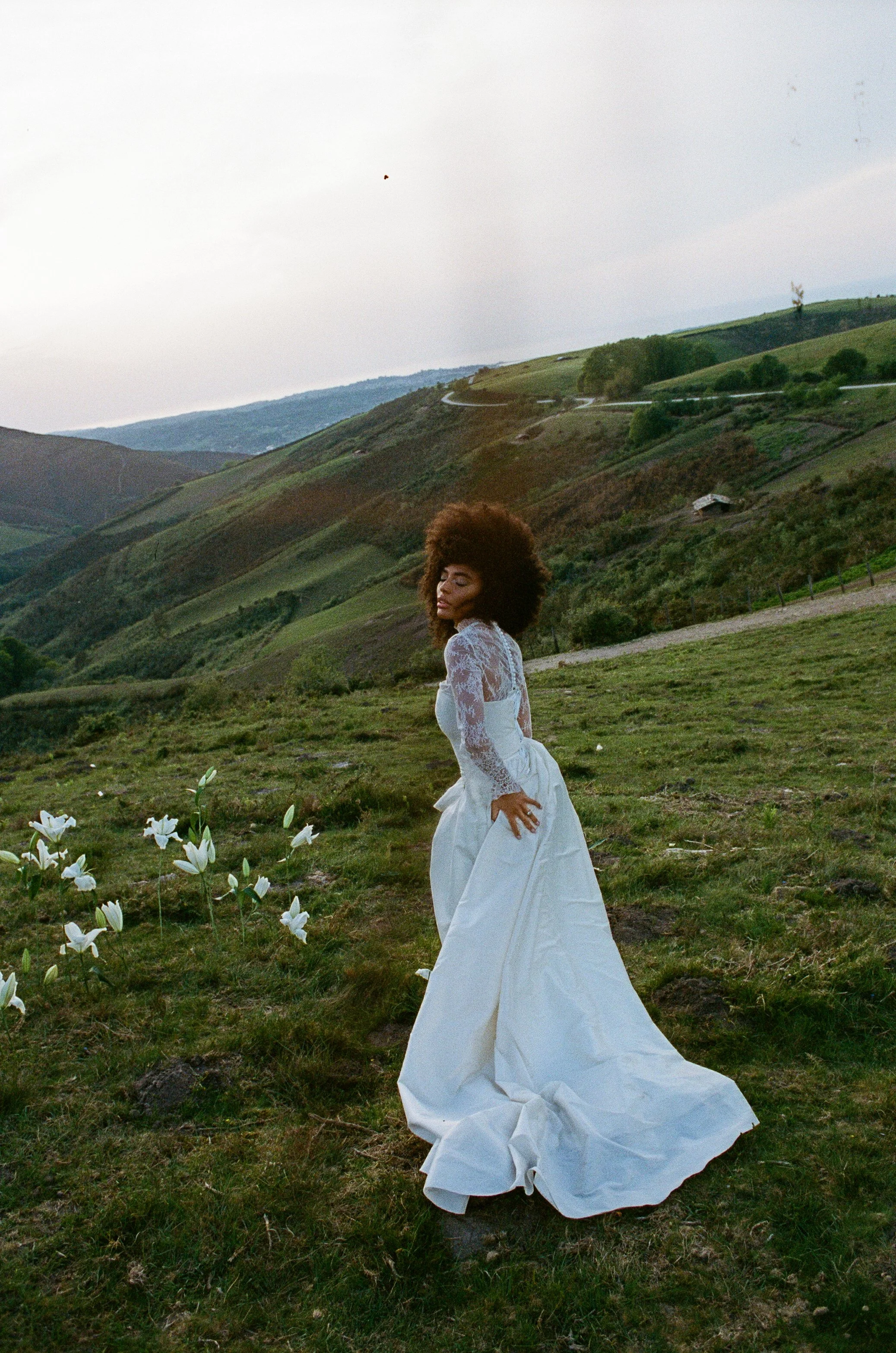 A woman dressed in a white gown standing outdoors on green grass with white flowers, with rolling hills and a cloudy sky in the background.