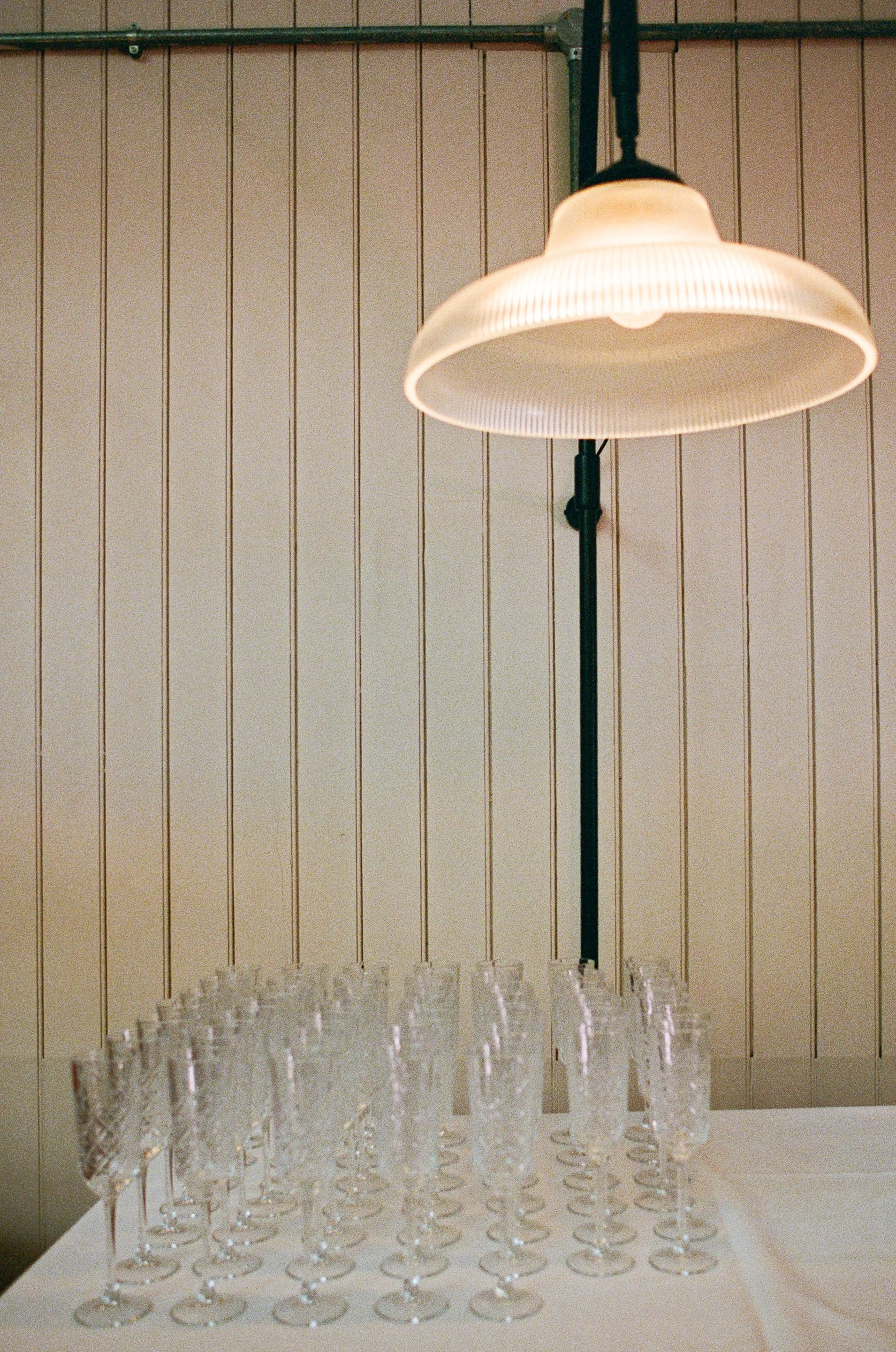 A large white table with numerous empty crystal champagne glasses arranged in rows. In the background, a wall with vertical panels and a hanging lamp above the table, casting light over the glasses.