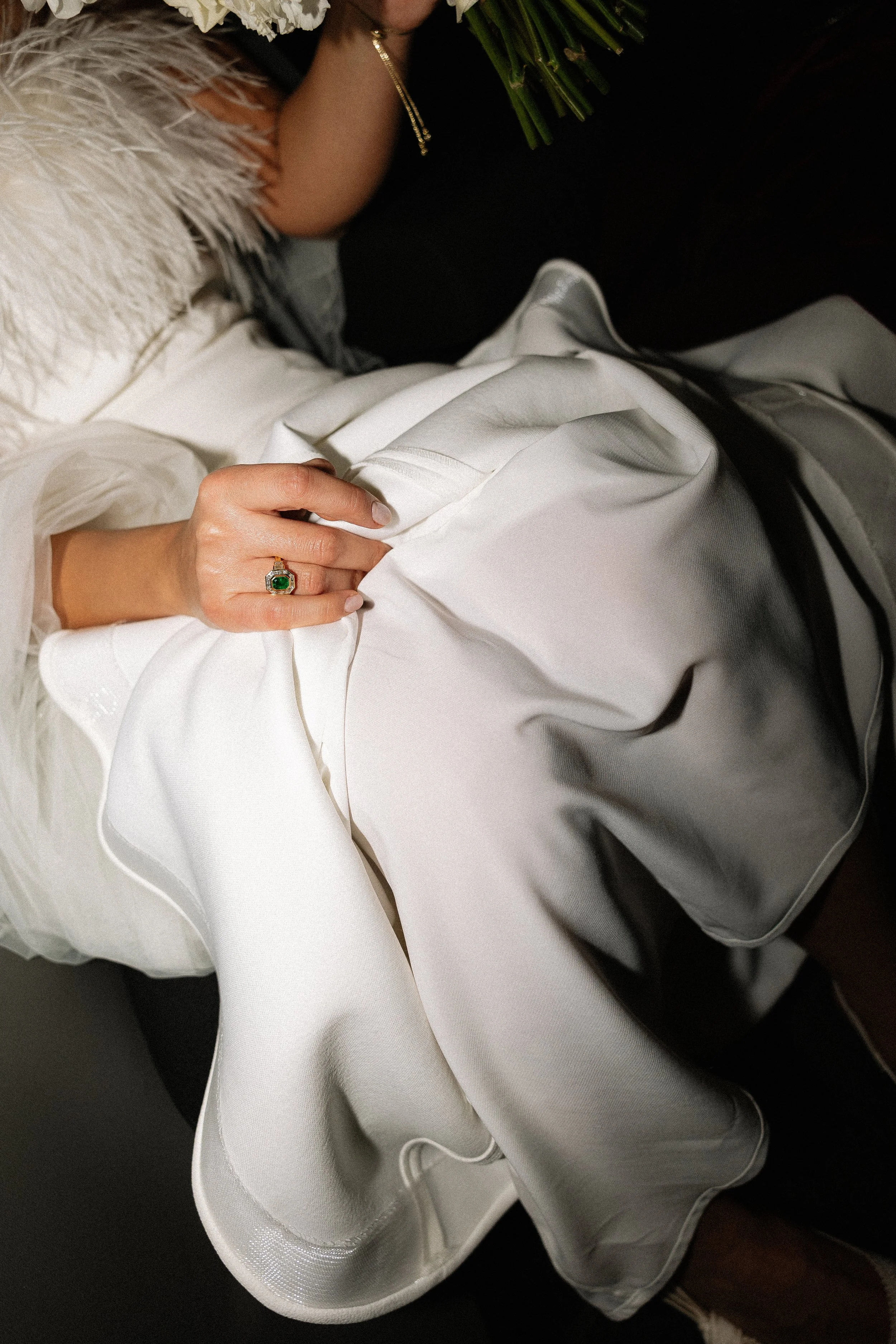 A woman lying on a bed holding a bouquet of green flowers, wearing a black dress, with a ring on her finger and a gold necklace, surrounded by white and beige pillows and satin sheets.