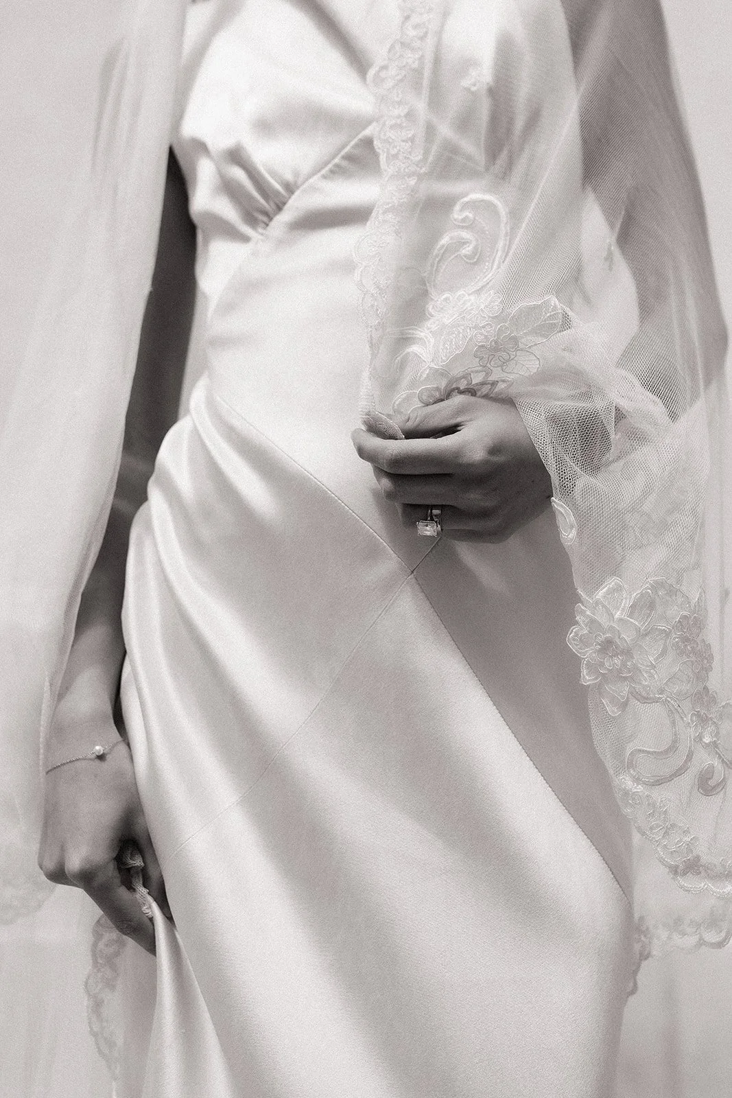 Close-up of a bride in a wedding gown with lace details, holding the dress with one hand, wearing a ring and bracelet.