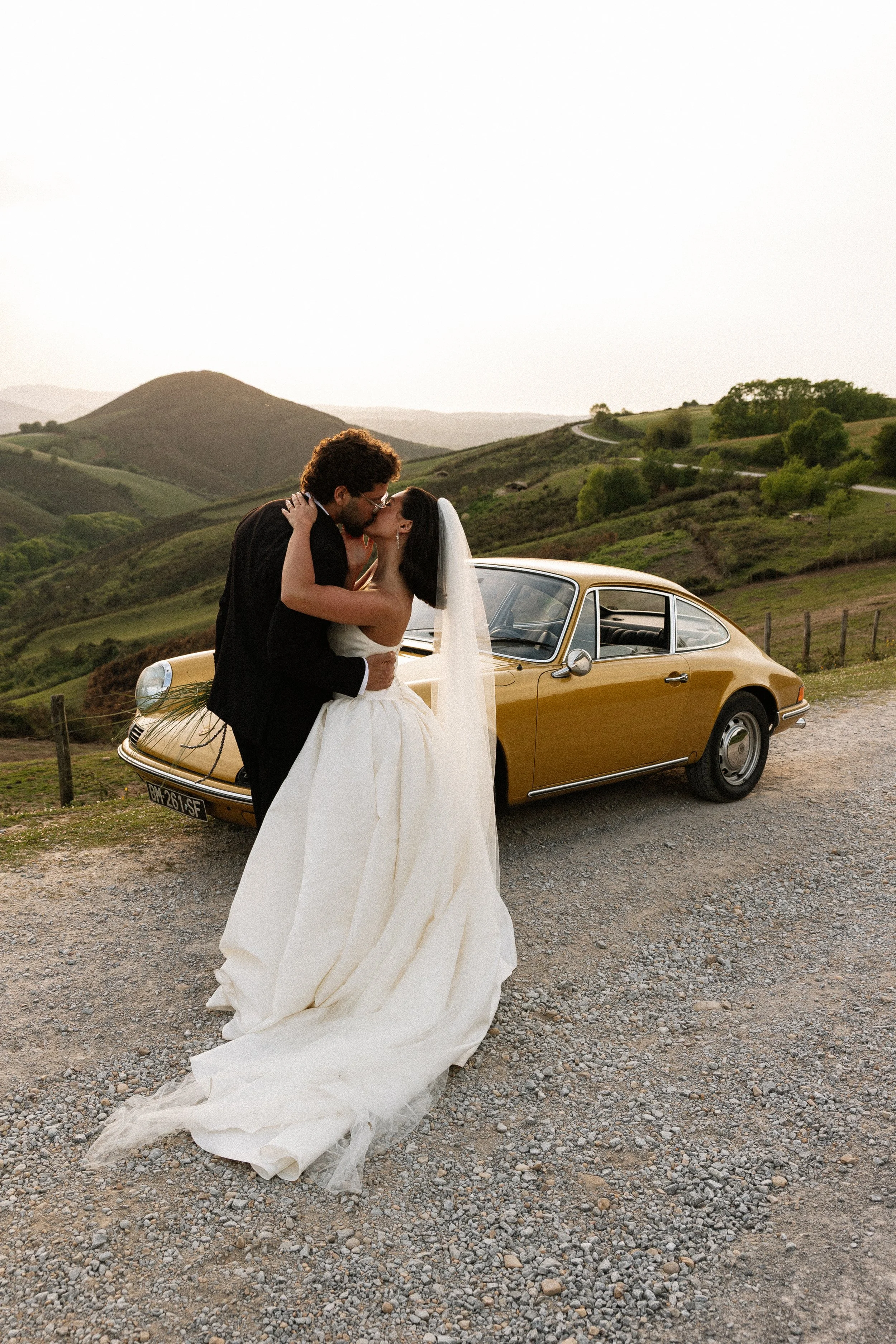 A bride and groom kissing in front of a yellow classic car with a countryside background of green rolling hills and a winding road during sunset.