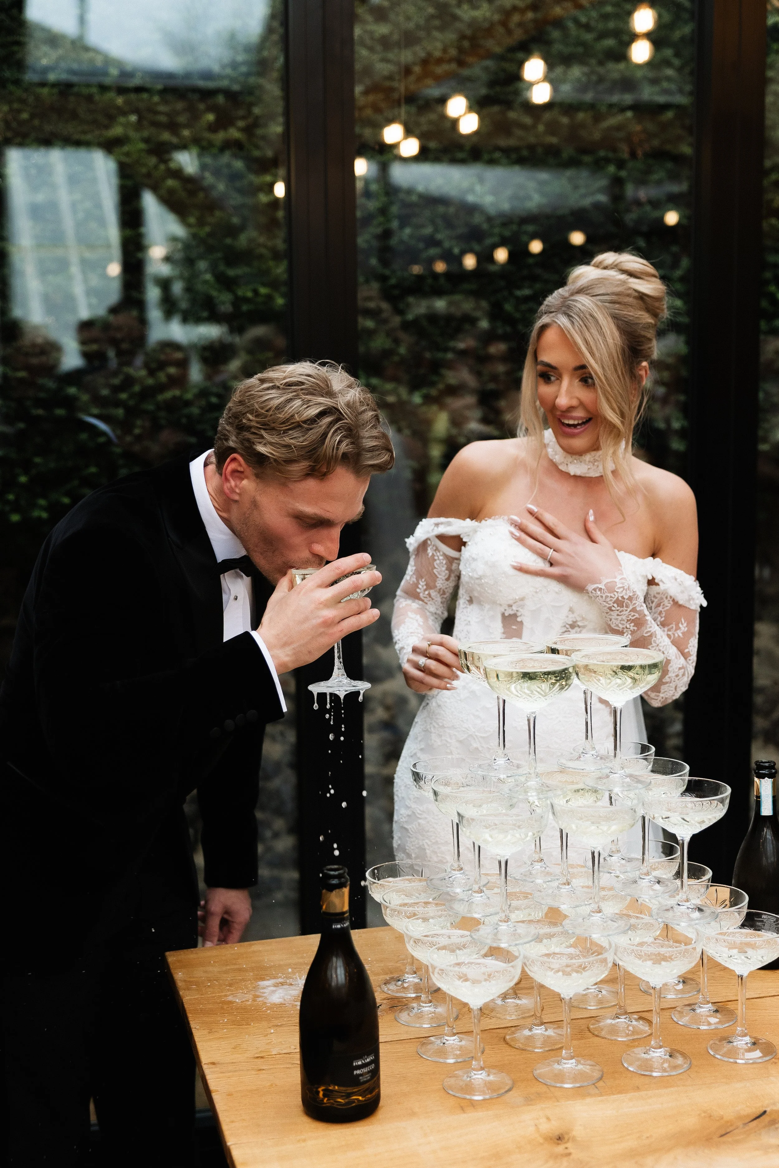 A bride and groom celebrate at a wedding reception with a champagne tower. The groom is in a tuxedo, drinking from a glass, while the bride in a lace wedding dress looks on smiling. The champagne tower is made of stacked glasses on a wooden table, and a bottle of prosecco is also on the table. The setting is indoors with large windows showing trees and warm lighting.