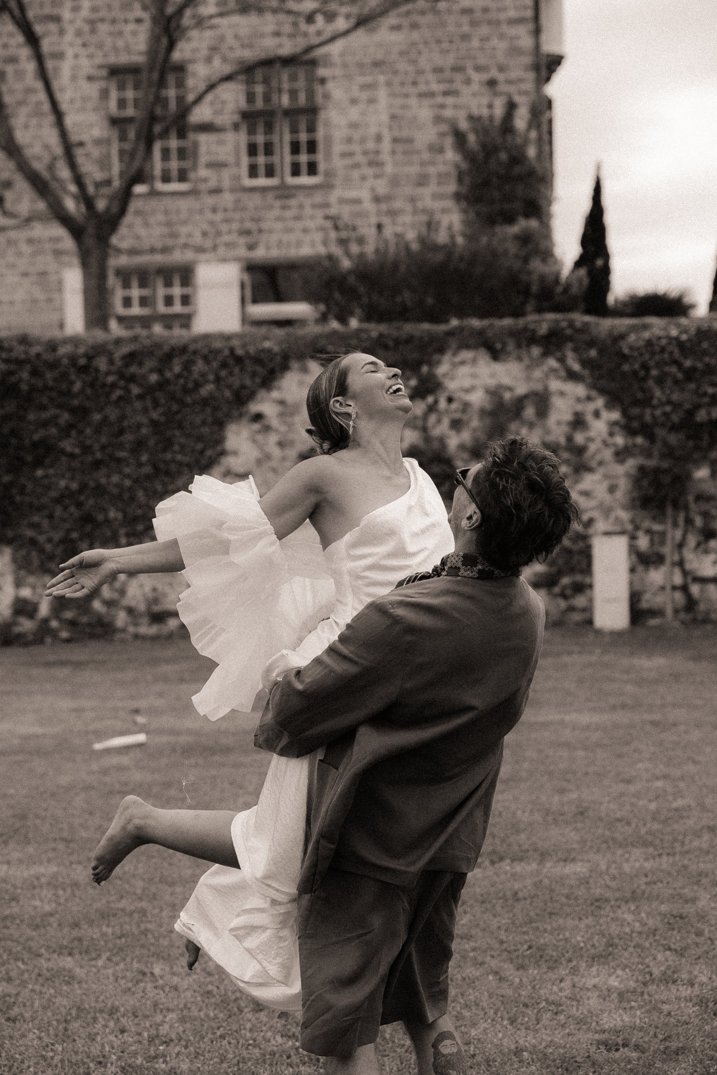 A woman in a white dress is being lifted by a man in sunglasses in an outdoor garden, both smiling and joyful, with a stone building and trees in the background.