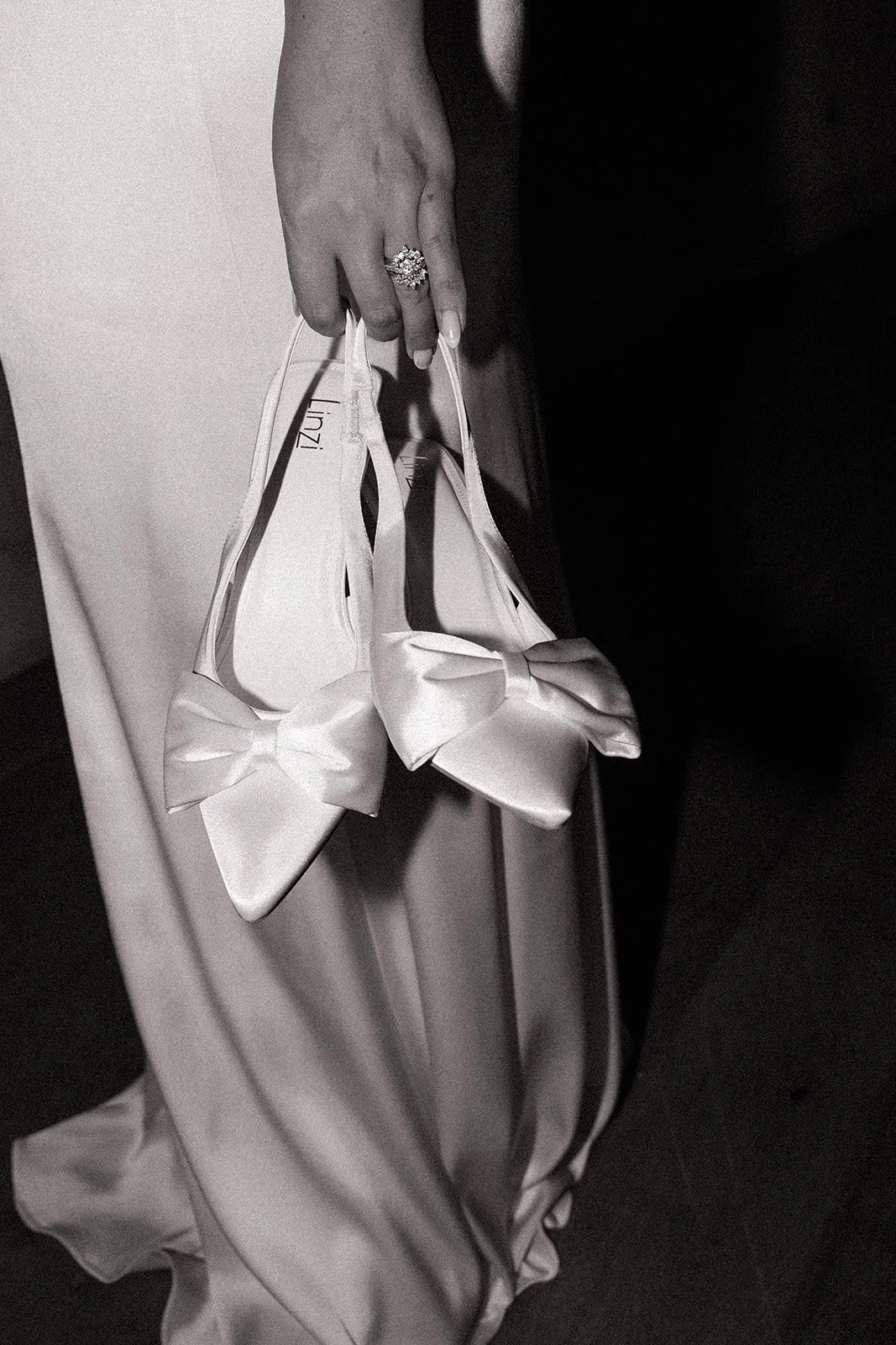 A person dressed in formal attire holding two satin high-heeled shoes with bow decorations, and wearing a large ring with a floral design.