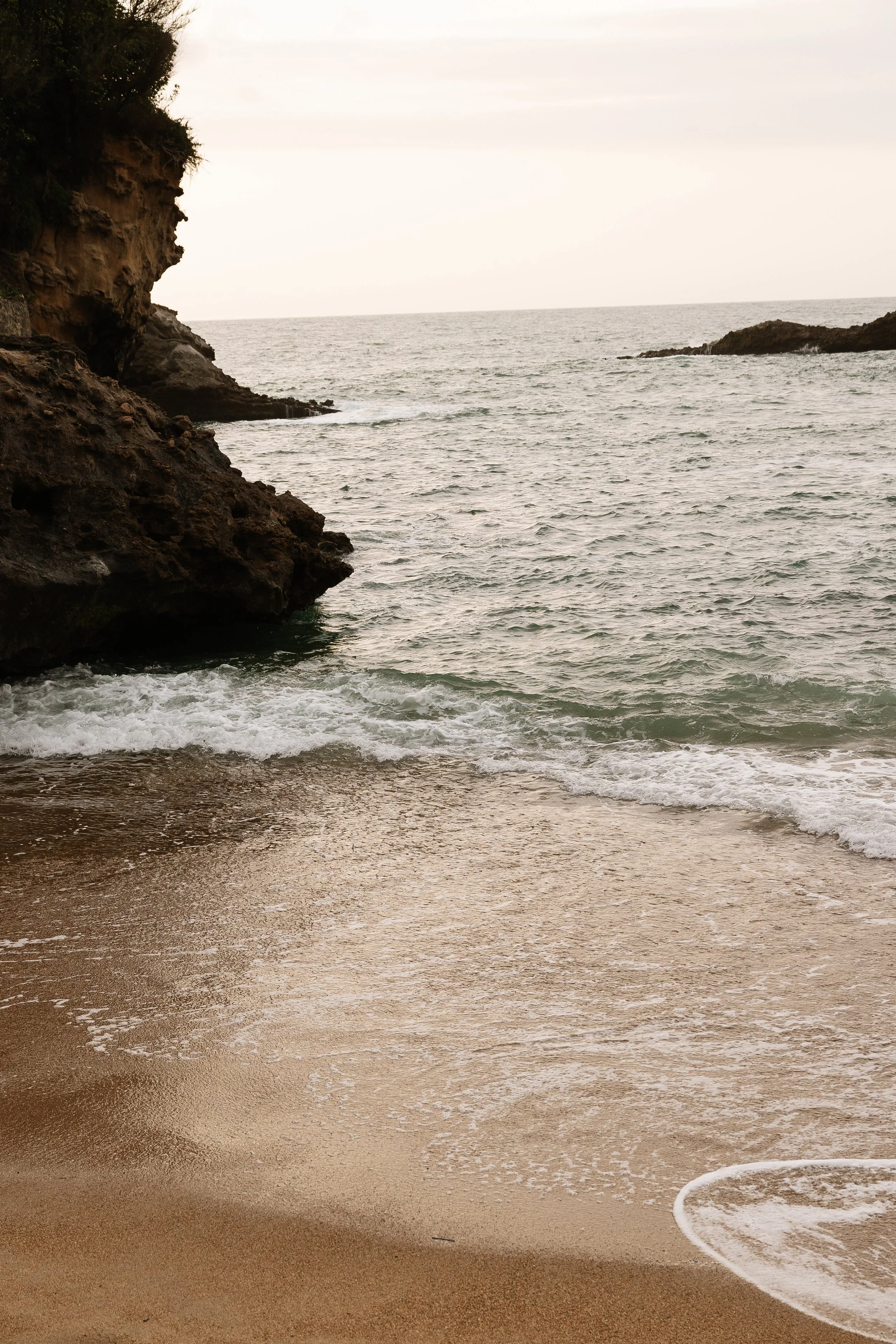 A sandy beach with gentle waves and rocky cliffs on the left side, under a cloudy sky.