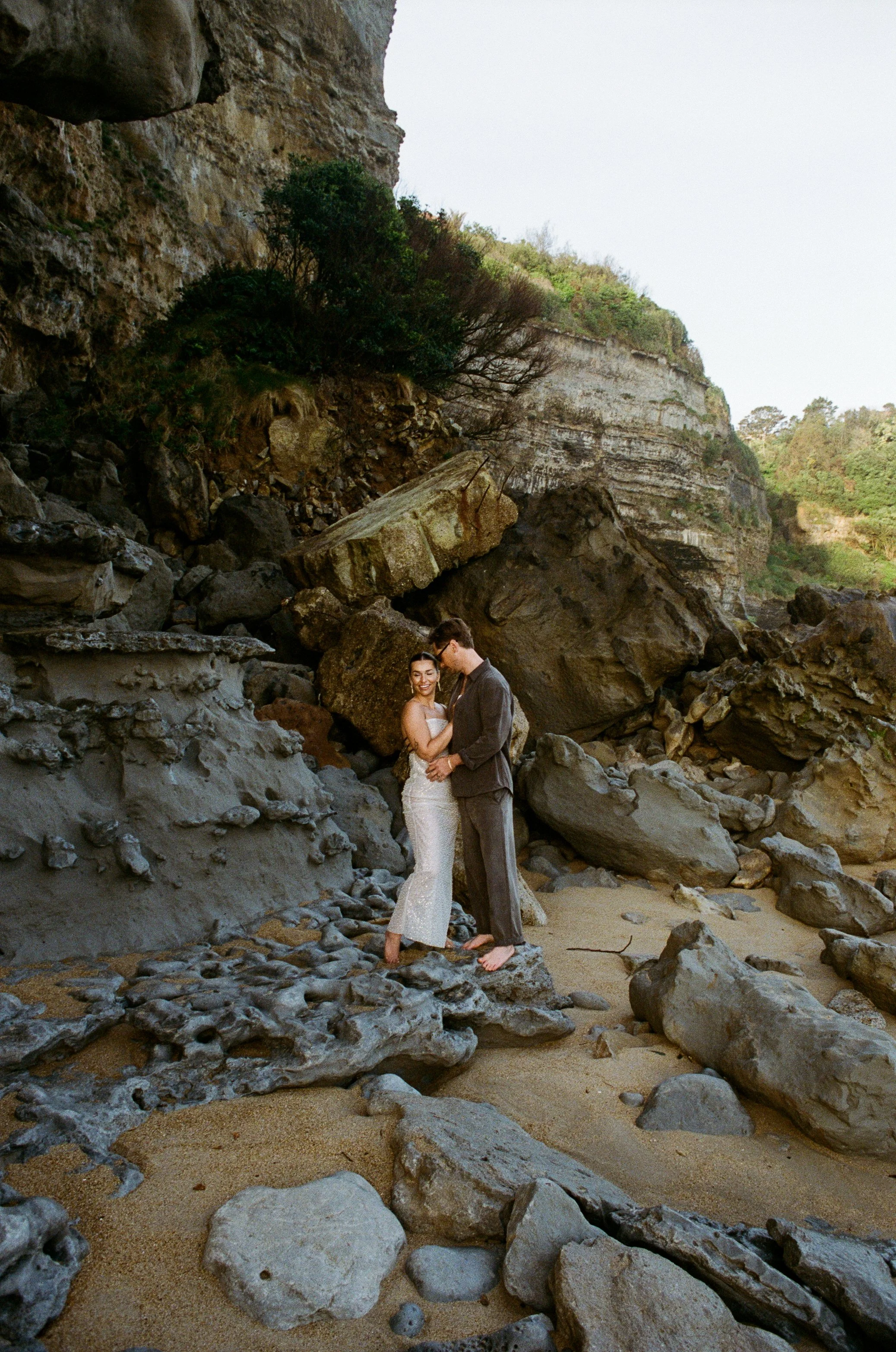 A couple in wedding attire standing on a rocky beach with cliffs in the background, holding hands and smiling at each other. Taken on 35mm analogue film.