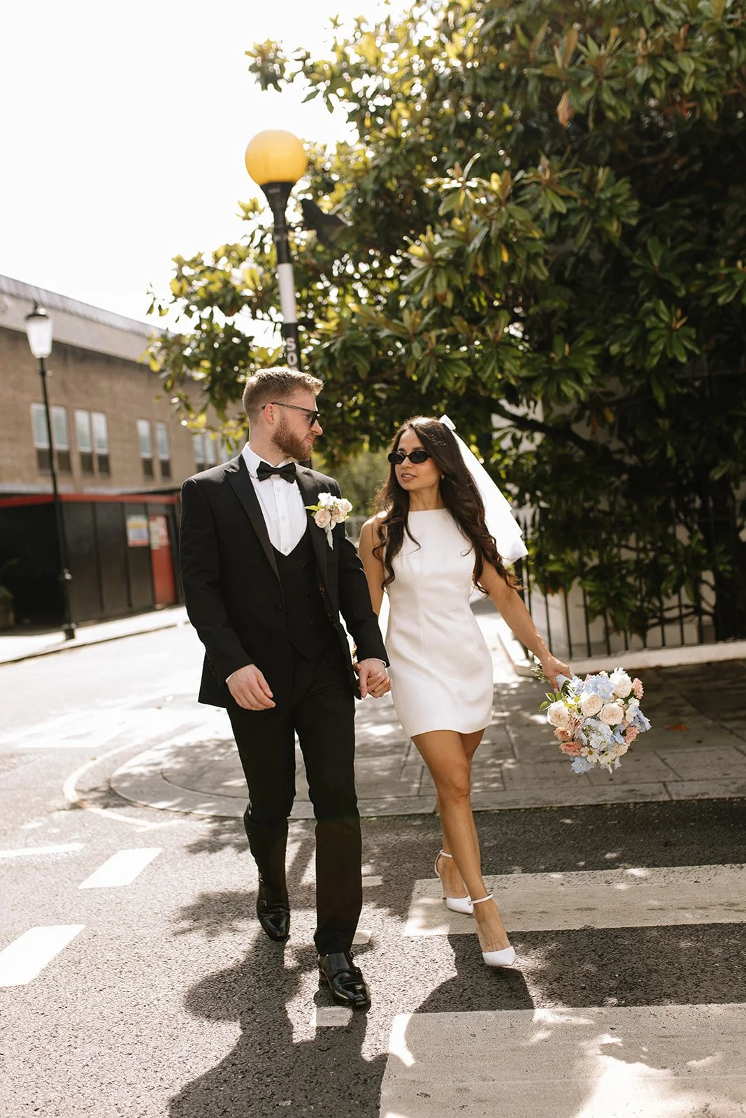 A bride and groom walking hand in hand across a crosswalk on a sunny day, with the bride holding a flower bouquet and wearing sunglasses.