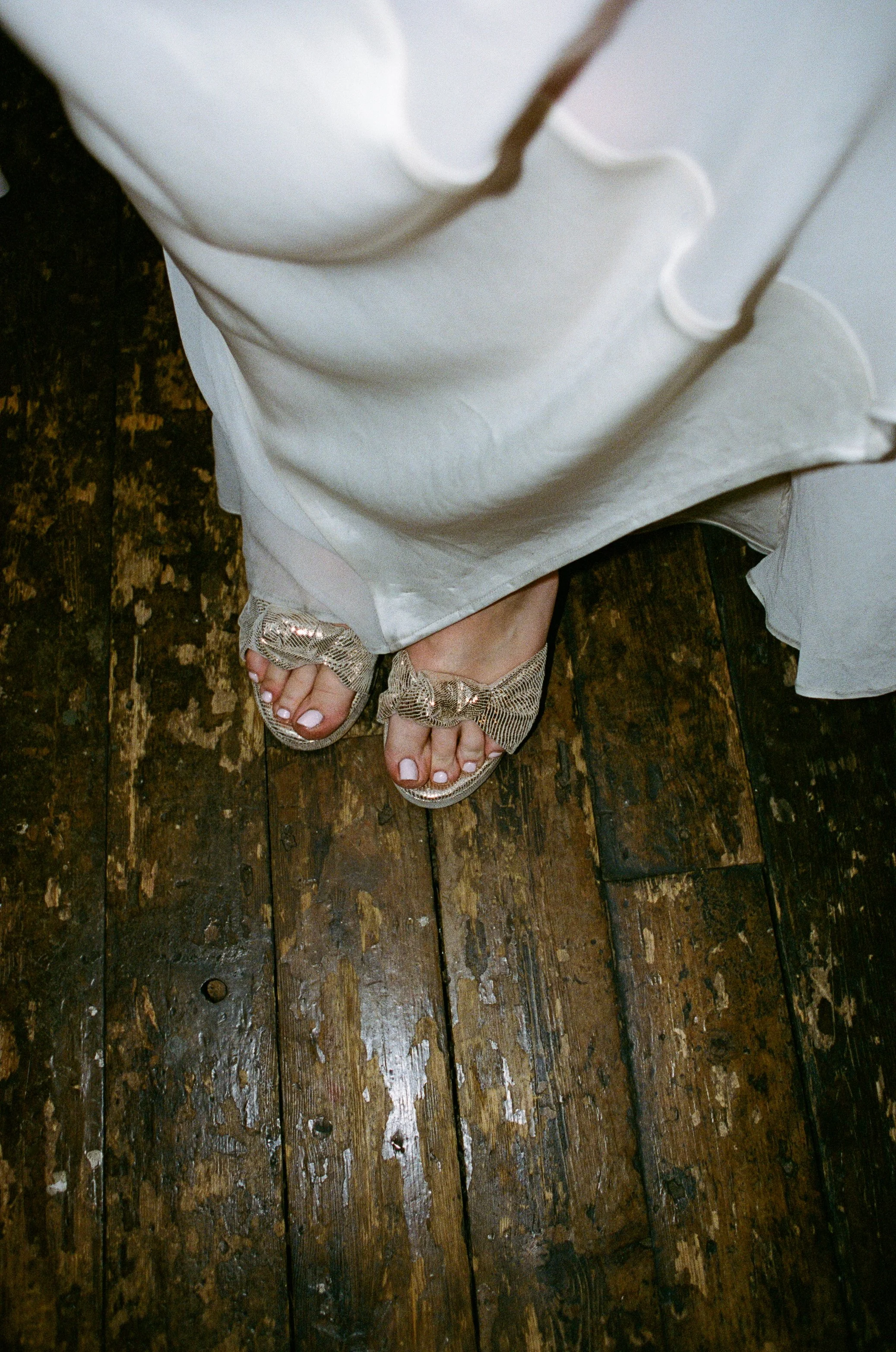 Top view of a person's feet wearing sandals with bows inside a white dress with lace details, standing on a dark wooden floor.