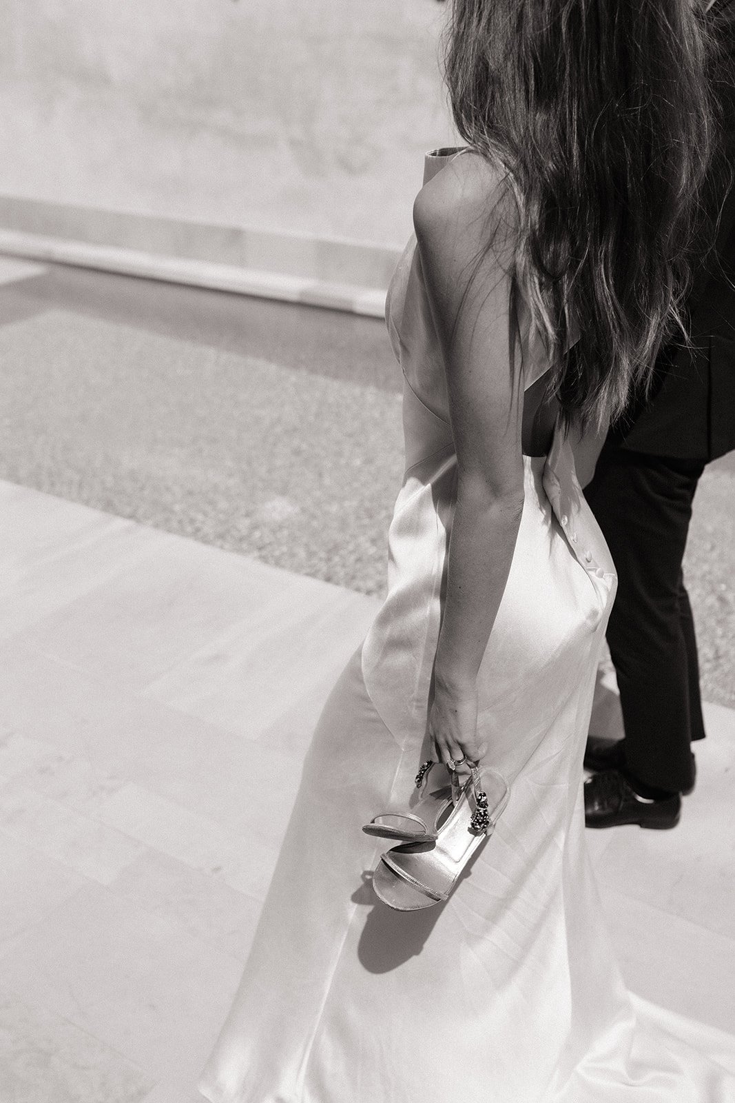 A woman wearing a satin wedding dress, holding a pair of high-heeled shoes, with another person partially visible in the background.