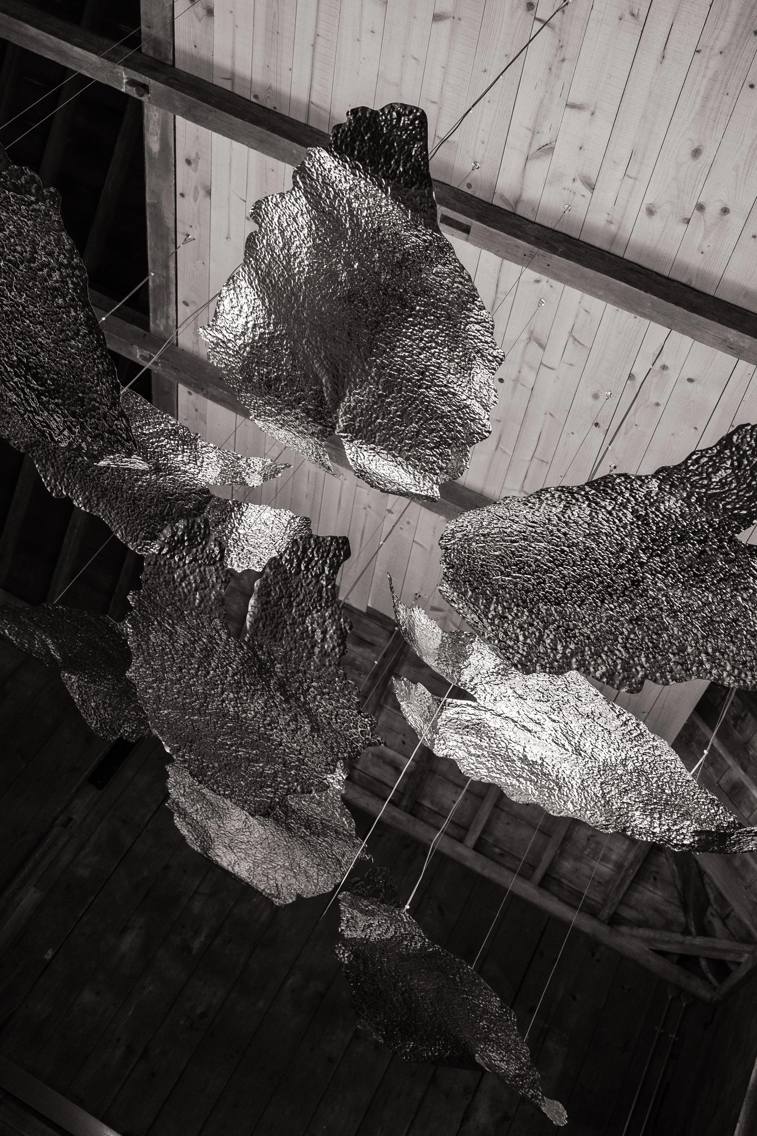 A black and silver abstract hanging sculpture with irregular, textured shapes, set against a wooden ceiling.