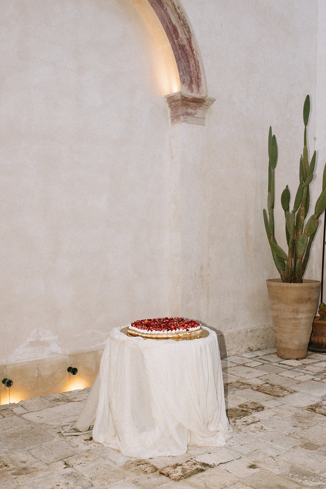 A pie with berries on top placed on a round table covered with a white cloth, in a room with stone flooring, minimalist wall, and a large potted cactus plant.
