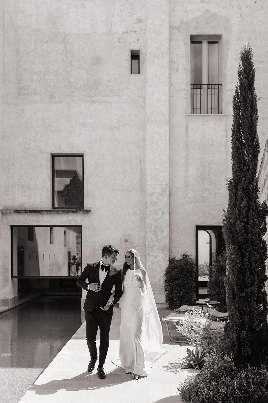 A bride and groom walking together outdoors in front of a modern building with windows, trees, and plants, dressed in wedding attire, smiling at each other.