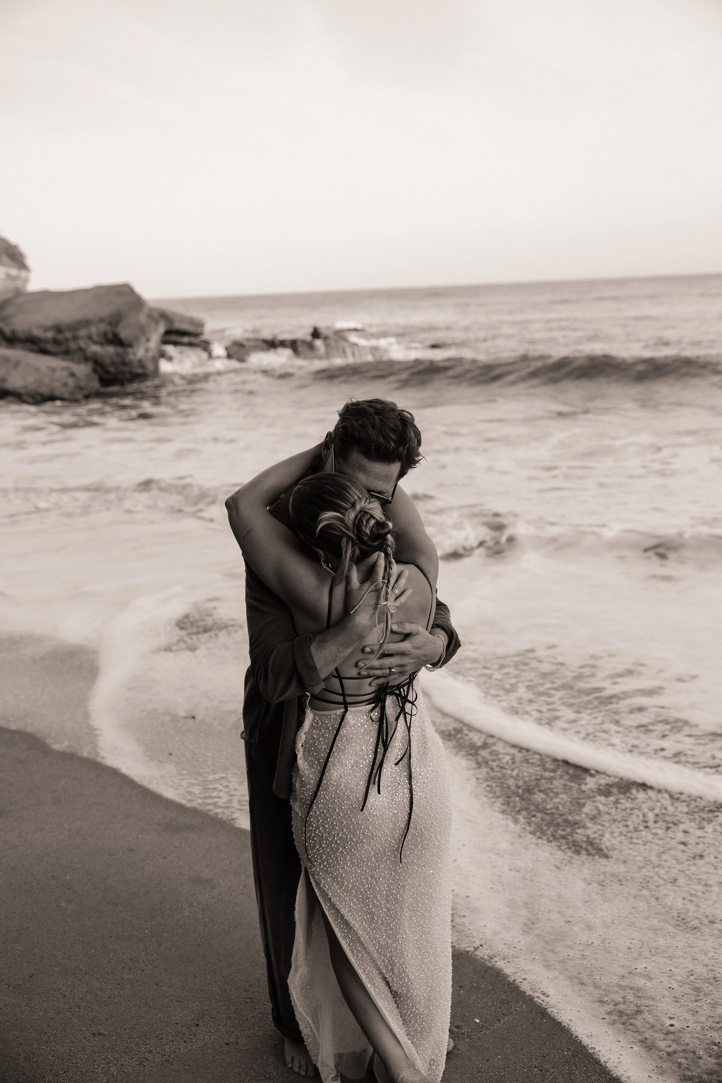 A couple embraces on a beach at sunset, with the woman wearing a sparkling gown and the man dressed formally, standing close to the shoreline with waves in the background.