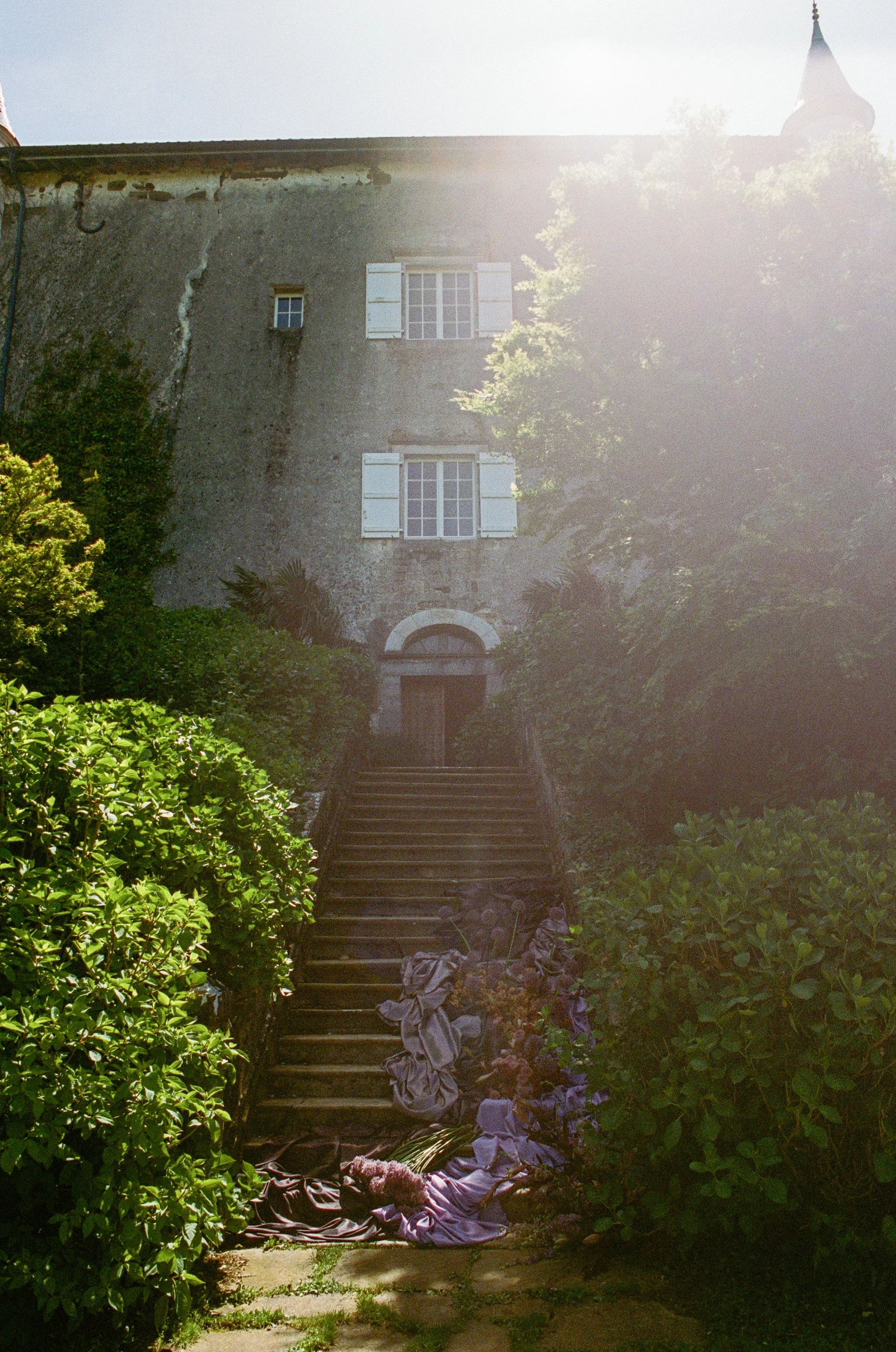 Stone building with three windows, white shutters, surrounded by greenery and a staircase leading to a wooden door, with clothes and laundry on the stairs in sunlight.