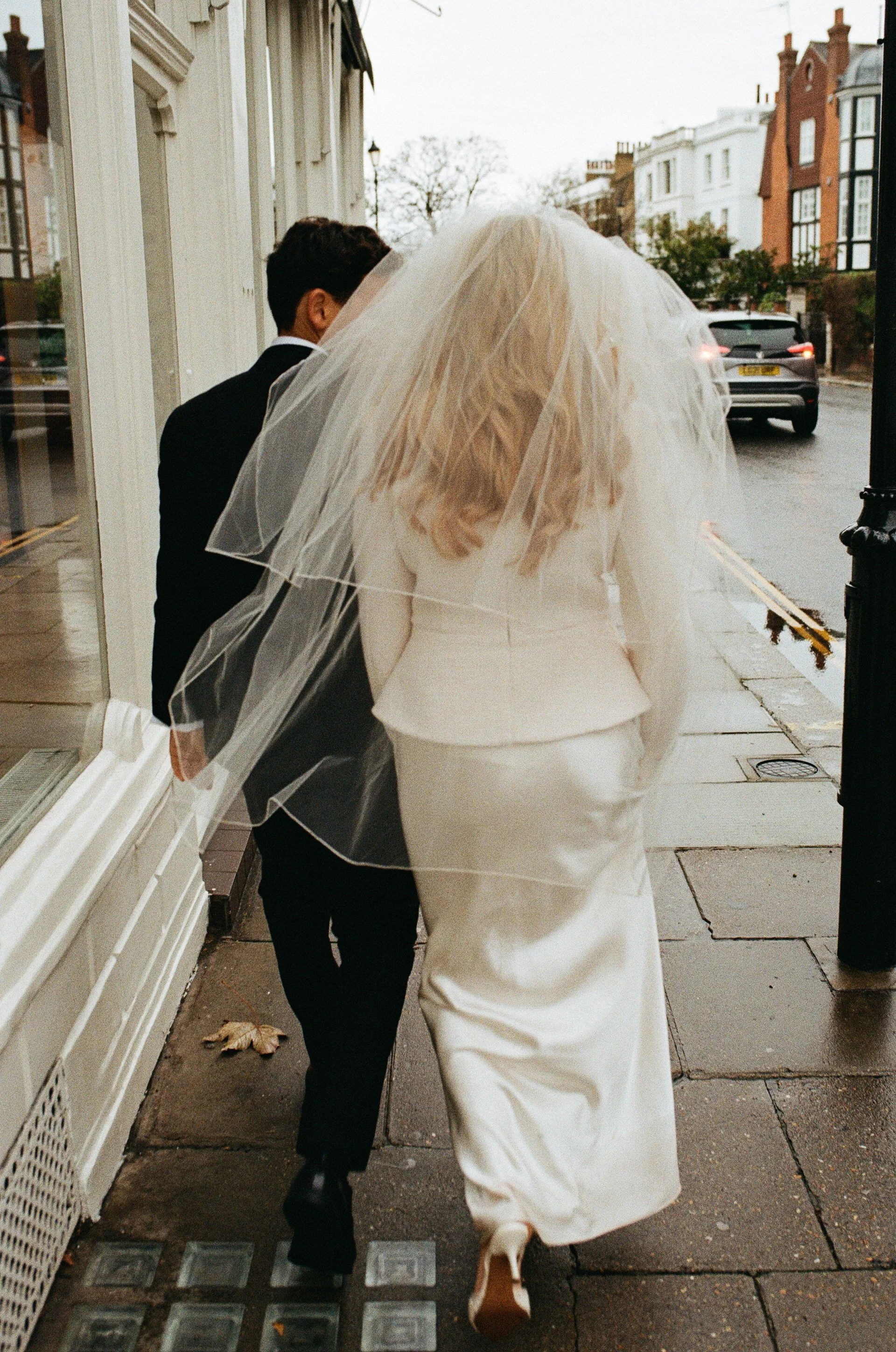 A bride and a young boy, possibly a ring bearer, walking together on a city sidewalk on a cloudy day. The bride is wearing a white wedding gown and veil, and the boy is dressed in a black suit. They are walking past a storefront window with reflections of the street and cars, with buildings and bare trees in the background.