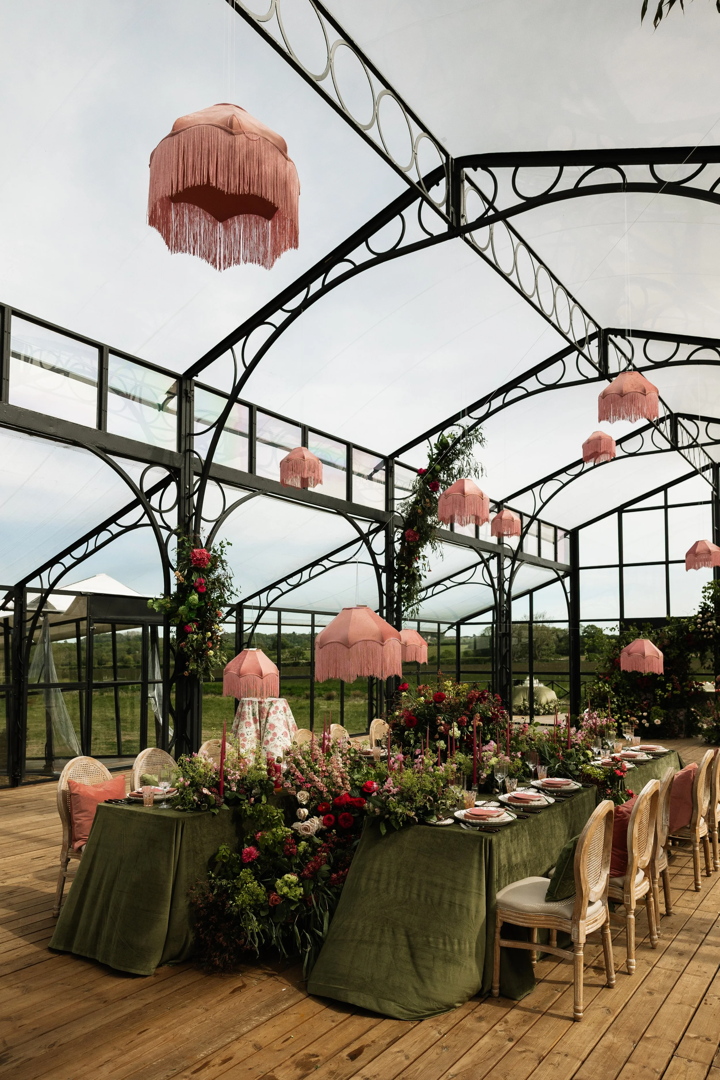 A decorated outdoor event space with a long table covered with a green cloth, set for a meal with plates, glasses, and floral centerpieces. Pink fringe umbrellas hang from a metal framework overhead, and the space features floral arrangements and greenery, with a backdrop of open fields and cloudy sky.