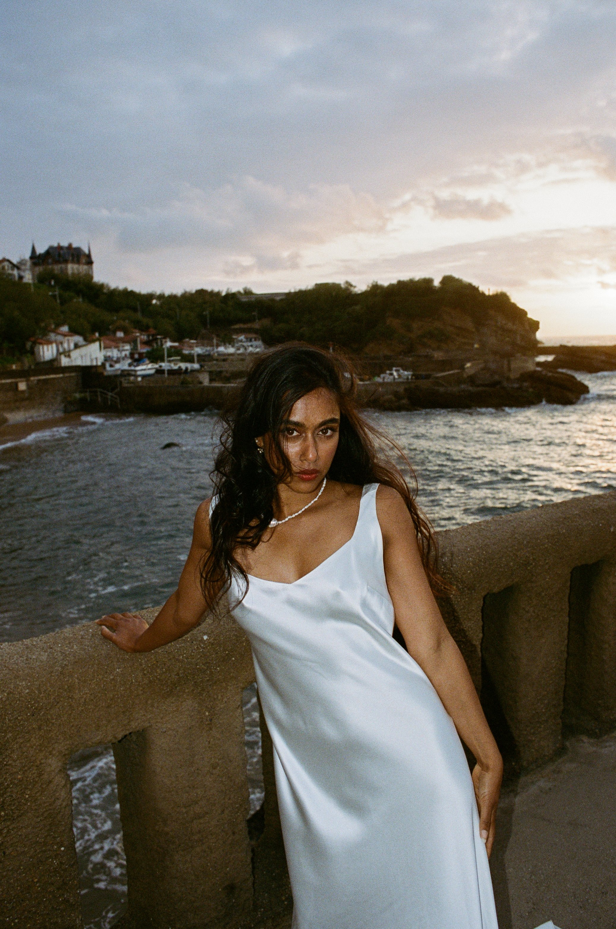 A woman in a white dress standing near a stone railing by the ocean at sunset, with a coastal town and a hilltop mansion in the background. Taken on 35mm analogue film.