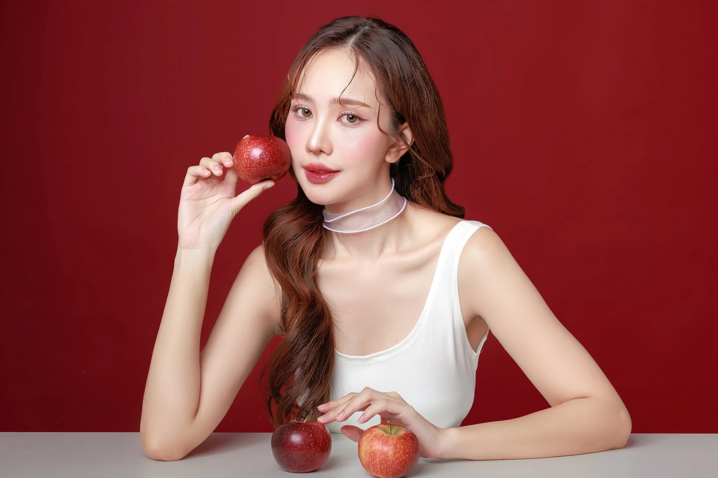 A young woman with long, wavy brown hair, wearing a white sleeveless top and a layered choker necklace, holds a red apple near her face while sitting at a table with two other apples in front of her against a red background.