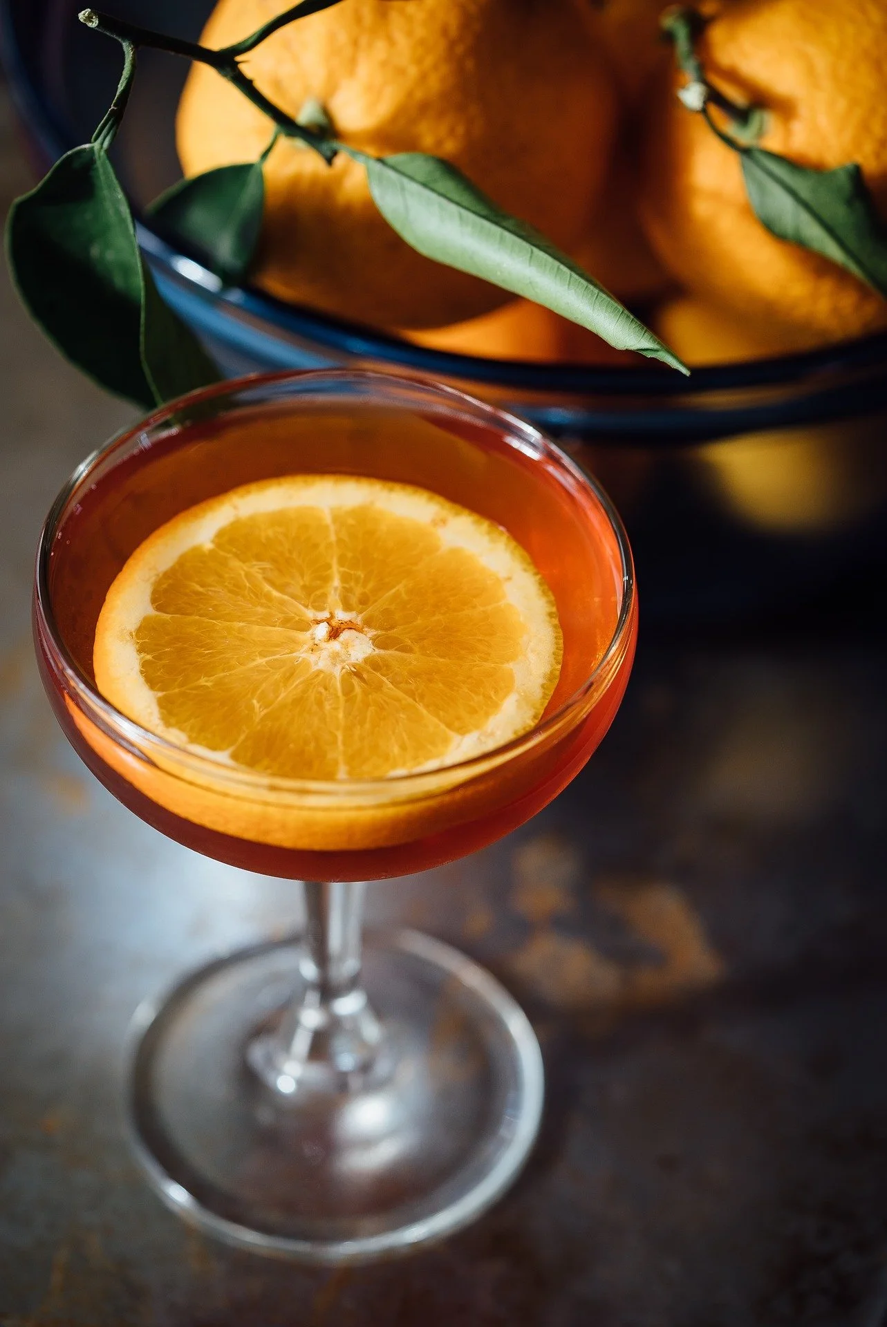 A cocktail glass with an orange slice inside, placed on a dark surface with a bowl of oranges and green leaves in the background.