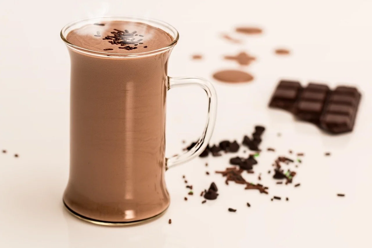 A glass mug filled with hot chocolate, topped with chocolate sprinkles, with chocolate pieces and sprinkles scattered around on a white surface.