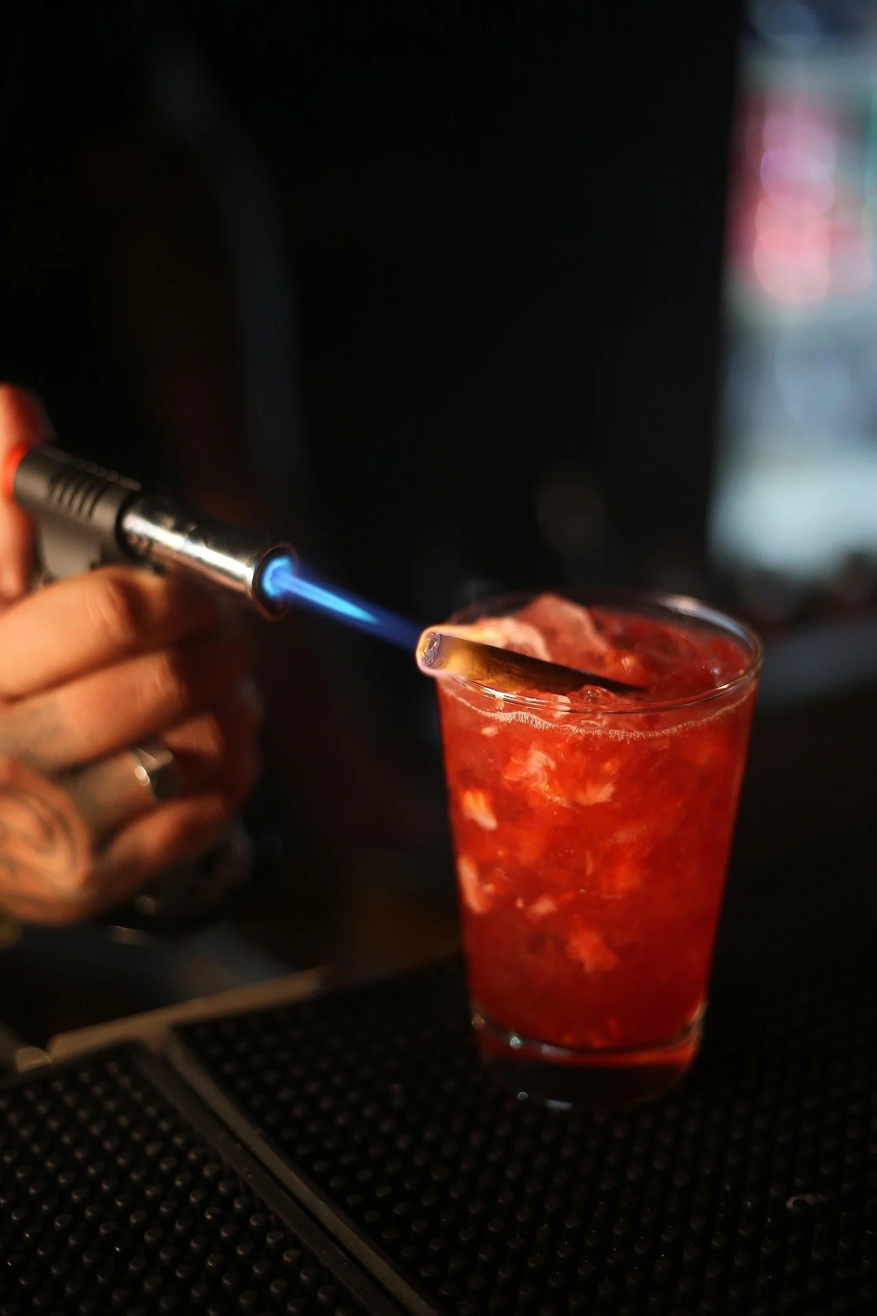 Person using a blowtorch to caramelize the top of a red cocktail drink in a tall glass.