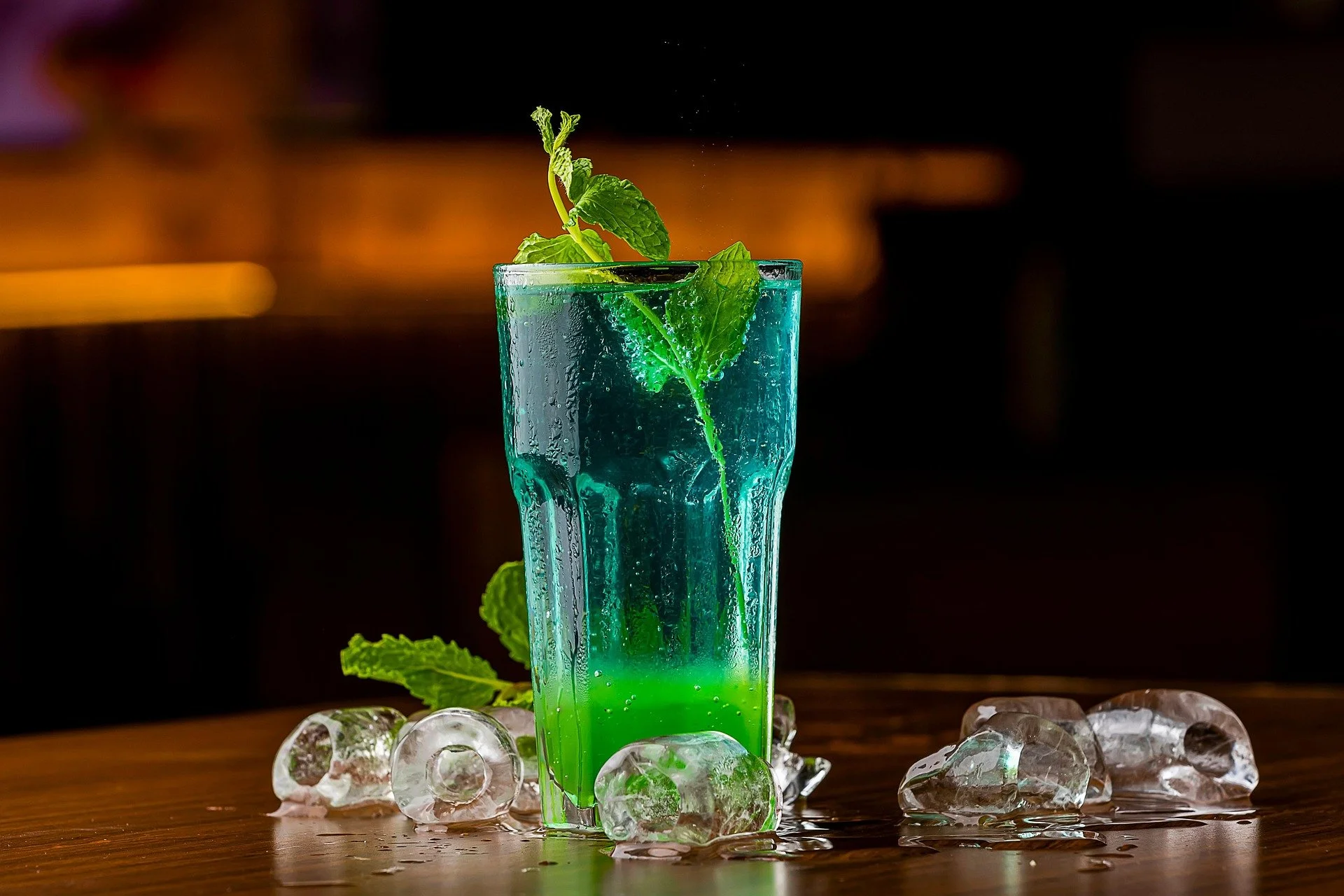 A colorful cocktail in a tall glass garnished with mint leaves, surrounded by melting ice cubes on a wooden surface.